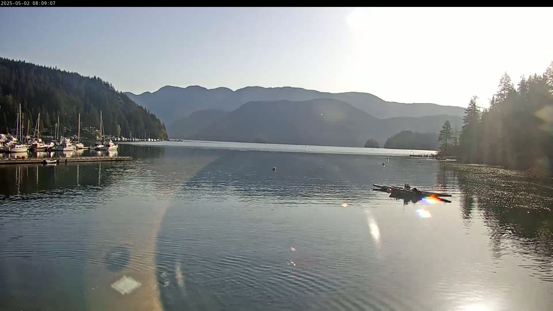 A calm body of water with docked boats and a small boat with people in it sits before a mountain range under a clear sky.