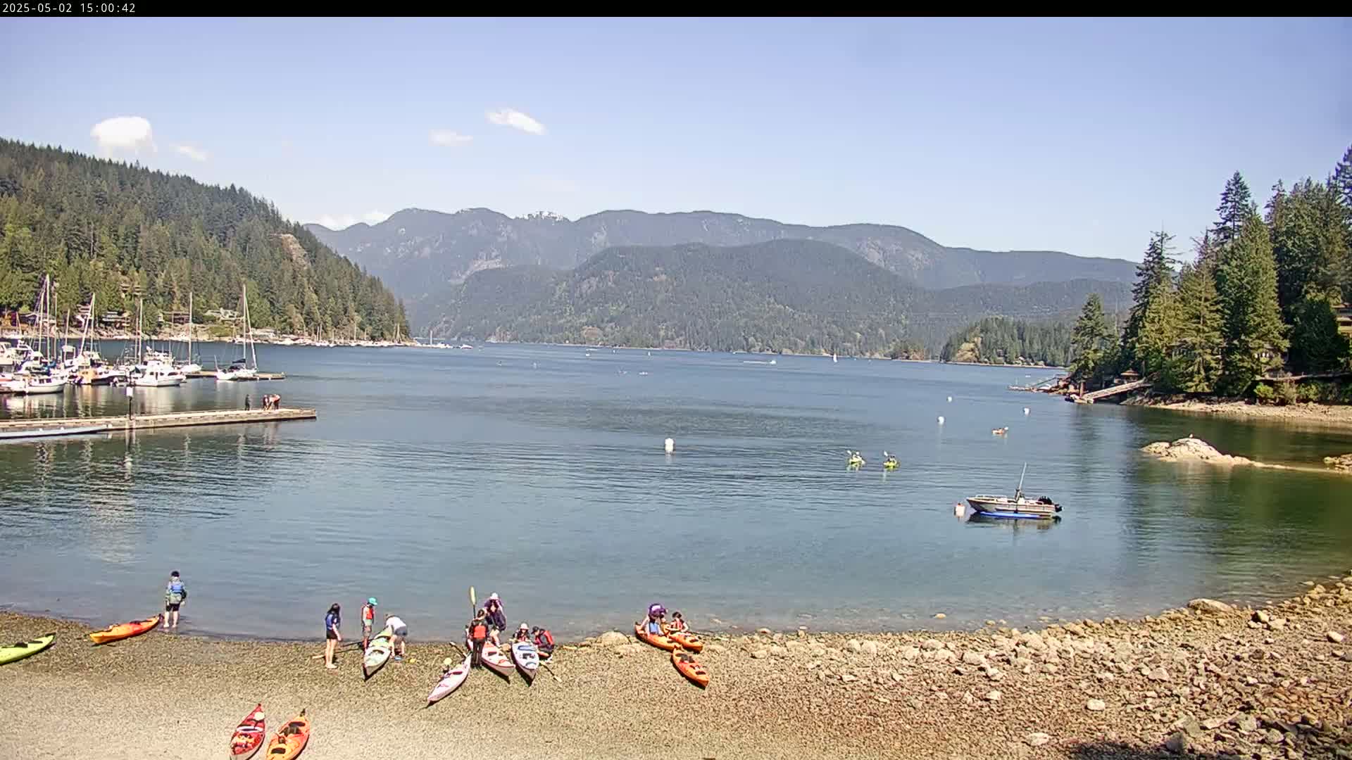 A calm bay with a rocky shore, several kayaks and people on the beach, a motorboat in the water, and numerous sailboats in a marina across the bay, all under a sunny sky against a mountainous backdrop.