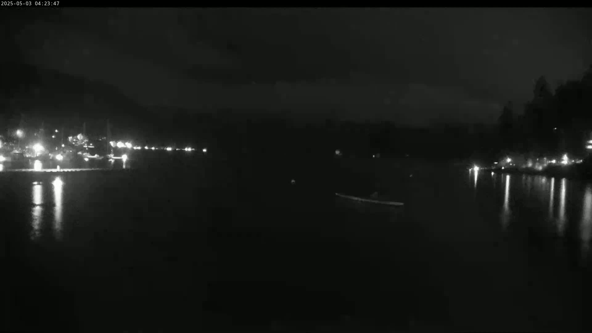A dark nighttime scene shows a body of water reflecting lights from a distant shore, with a small object possibly a boat or person in the water.
