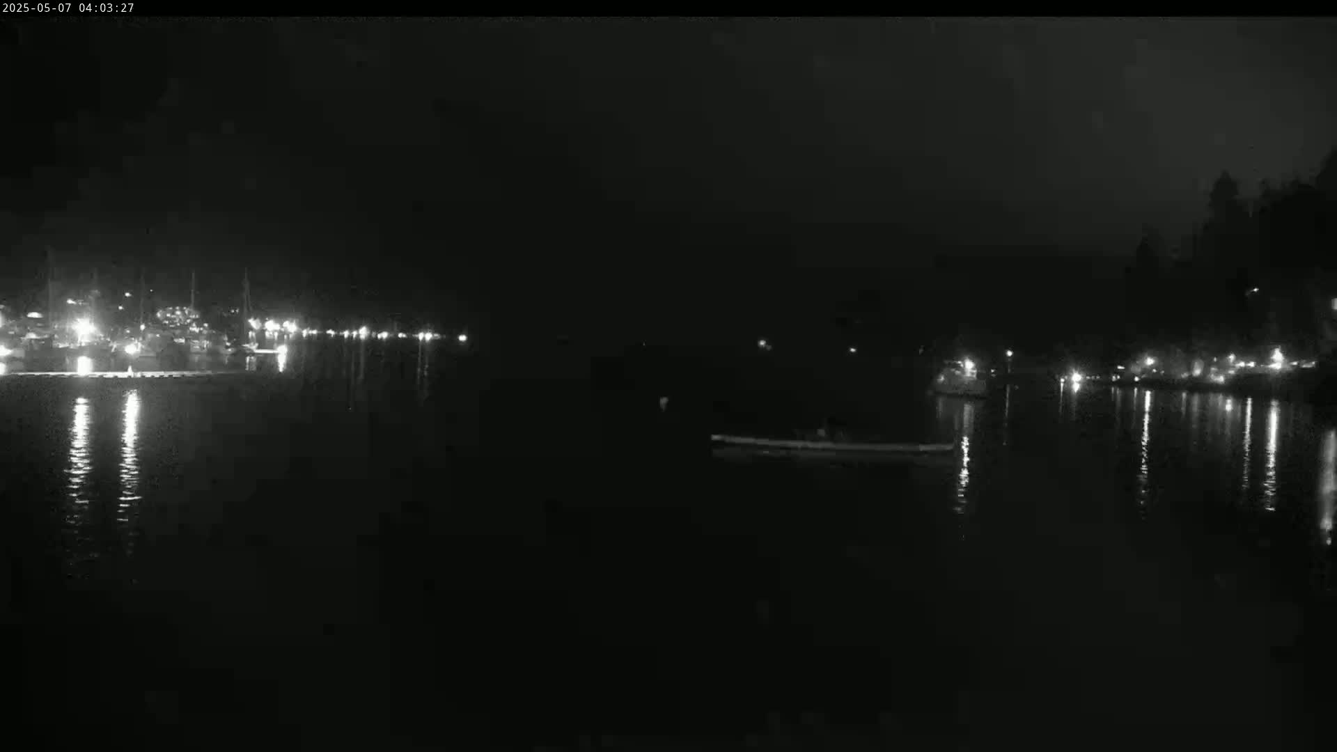 At night, a dark body of water reflects the lights from a distant shoreline and a few lights on the water.