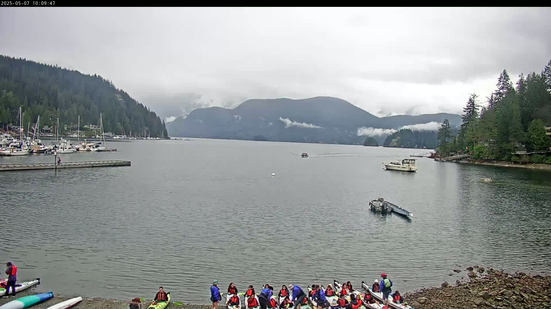 A group of people prepares kayaks on a rocky shore next to calm water, under an overcast sky, with boats moored in a marina and mountains in the background.