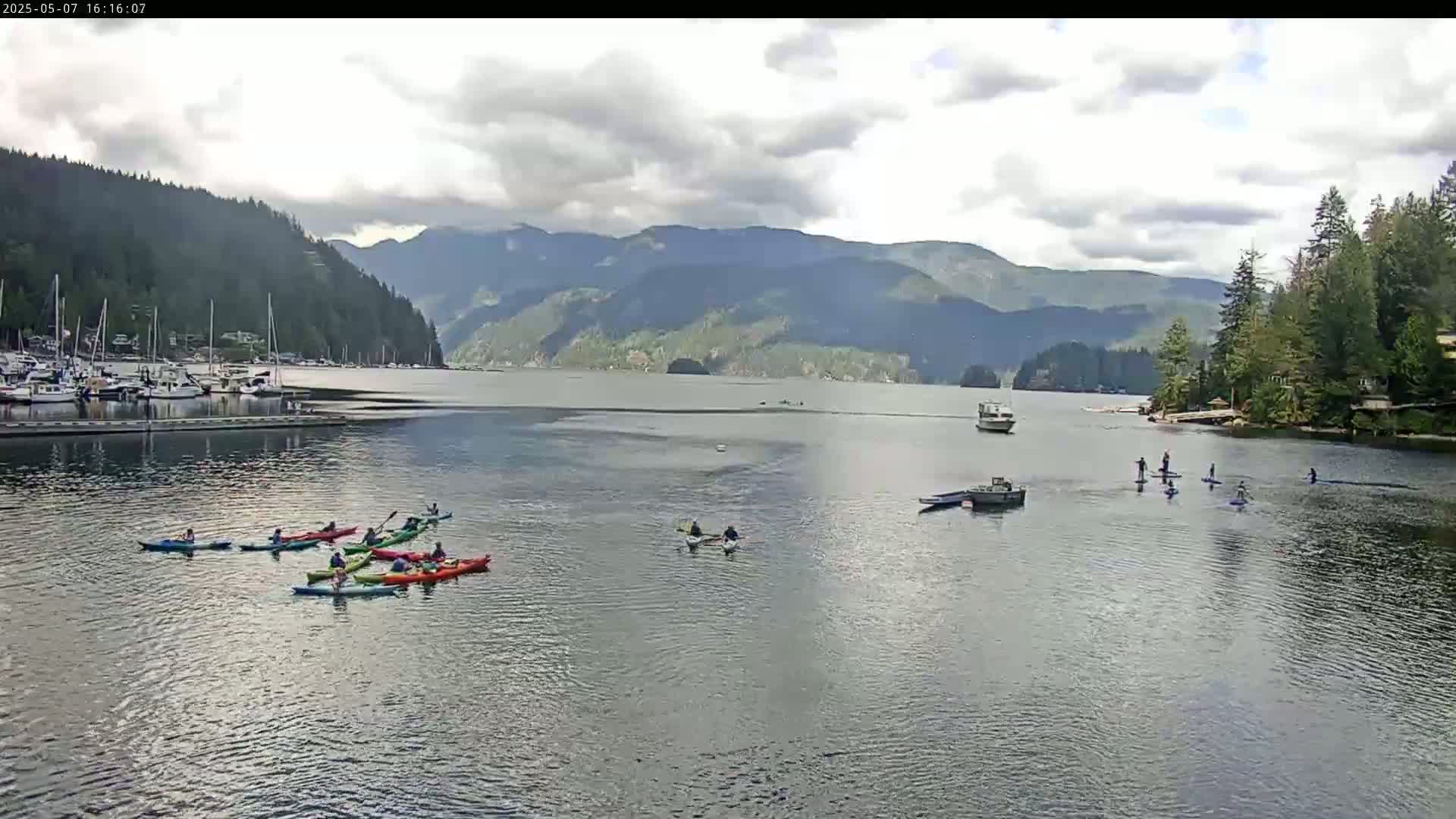 Kayakers and paddleboarders enjoy a calm, partly cloudy day on a bay surrounded by mountains and a marina.