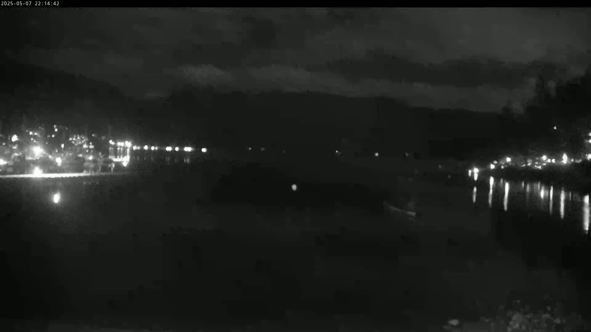 A nighttime, low-light view of a body of water reflecting the lights of a distant town under a mostly cloudy sky.