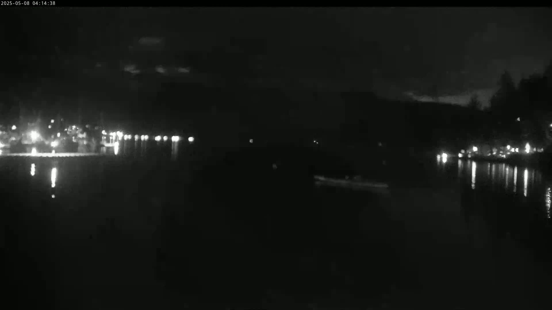 A dark, nighttime scene shows a body of water reflecting the lights from buildings and trees on the opposite shore under a mostly clear sky.