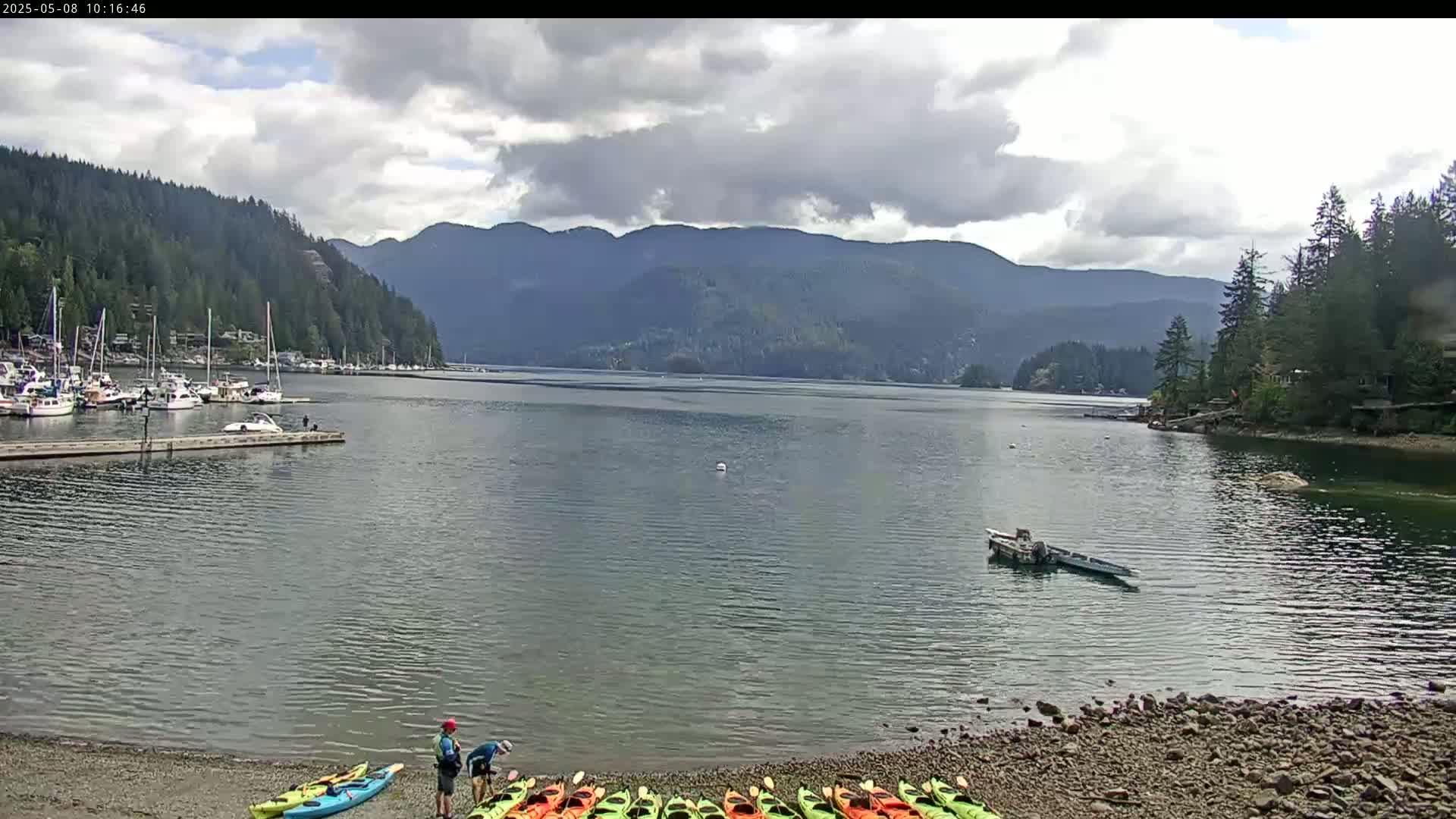 A calm bay with a marina full of boats, a rocky shore with kayaks, and mountains in the background under a partly cloudy sky.