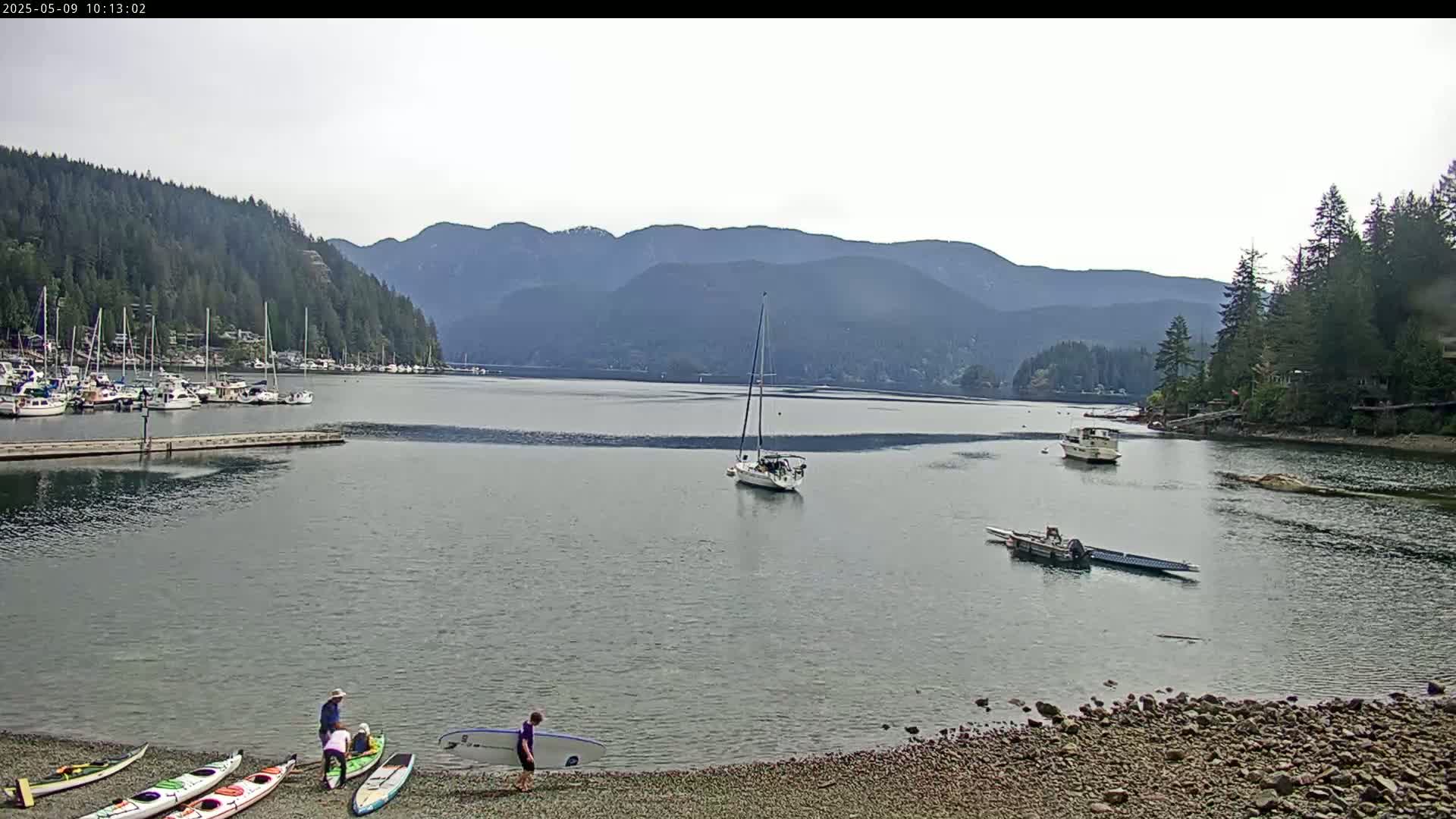 A calm bay, nestled between forested mountains under a mostly clear sky, features a marina, several boats at anchor, and people preparing kayaks and a surfboard on a rocky shore.