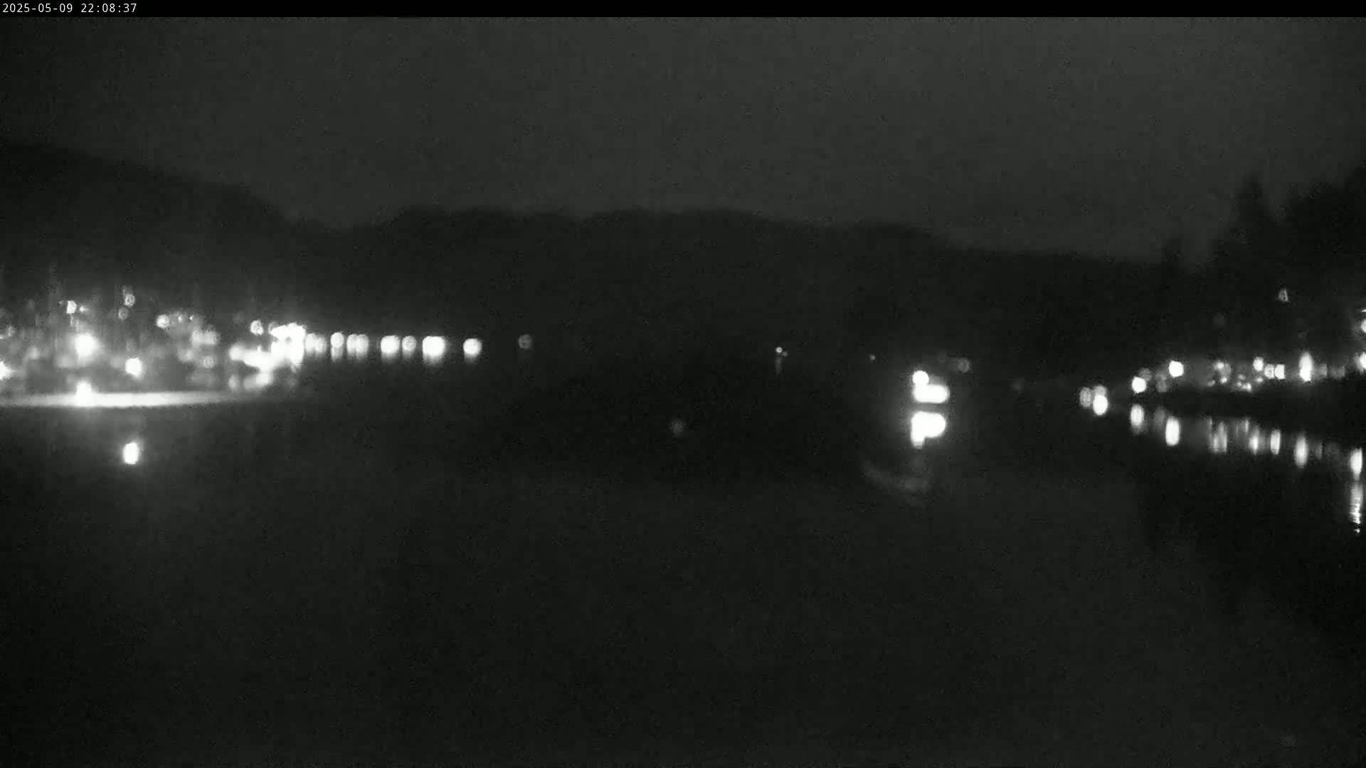 At night, blurry lights from buildings and their reflections are visible across a dark body of water, with a dark hill silhouetted in the background.