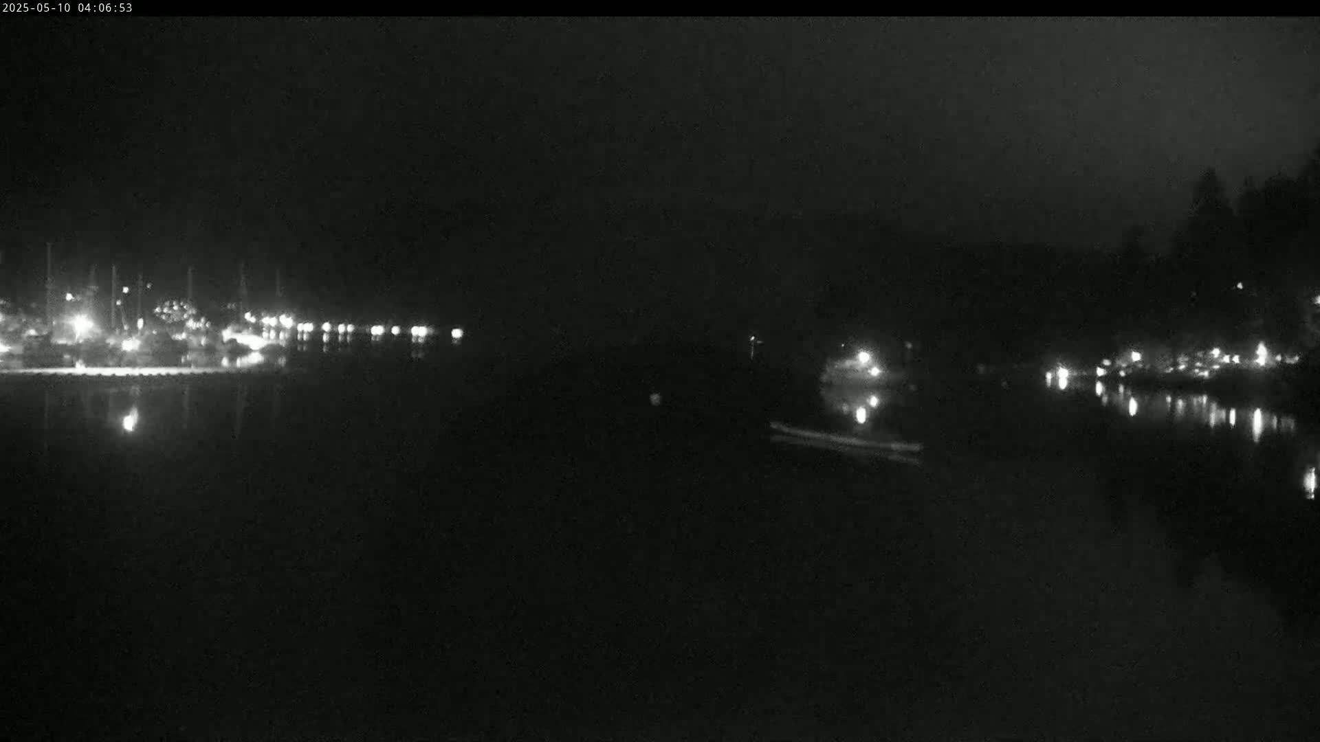 A dark, nighttime scene shows a body of water with faint lights reflecting on its surface, and a shoreline with sparsely scattered lights on either side.