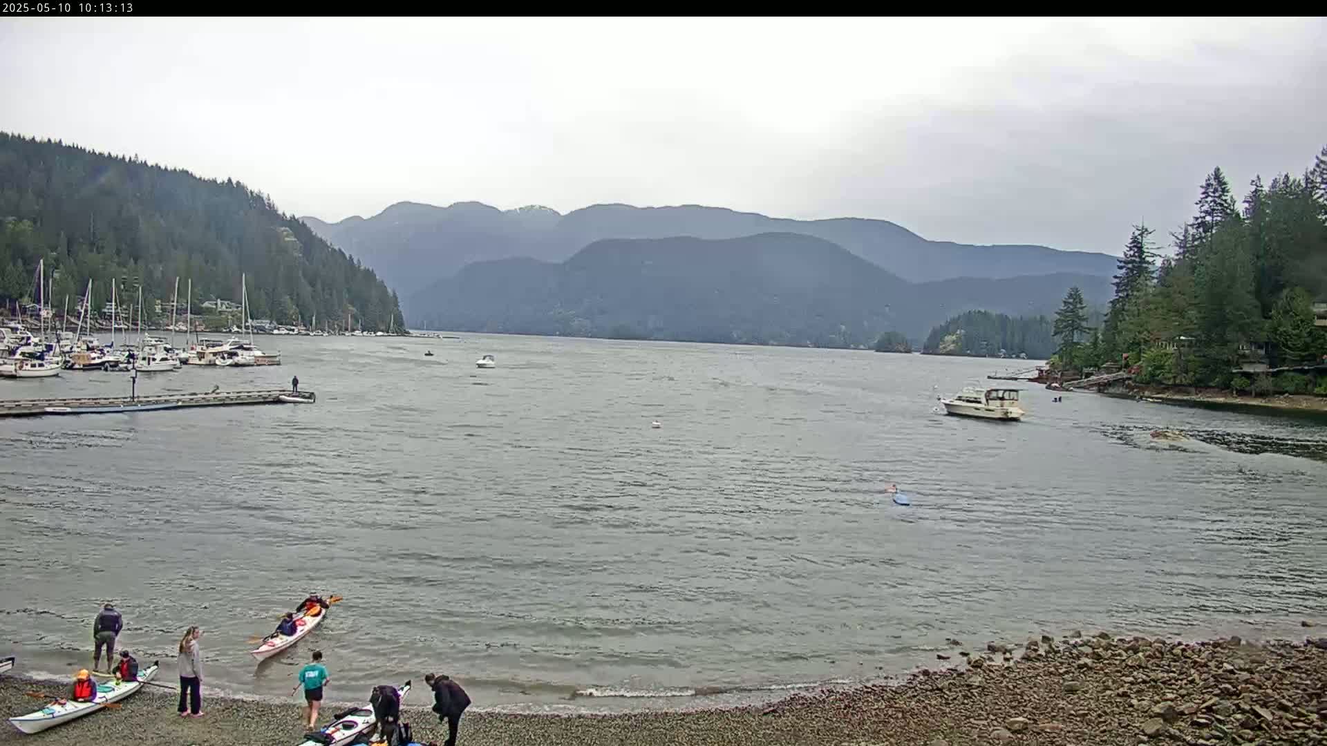 A calm, overcast day shows a bay with numerous boats docked, kayakers on the water and shore, and forested mountains in the background.