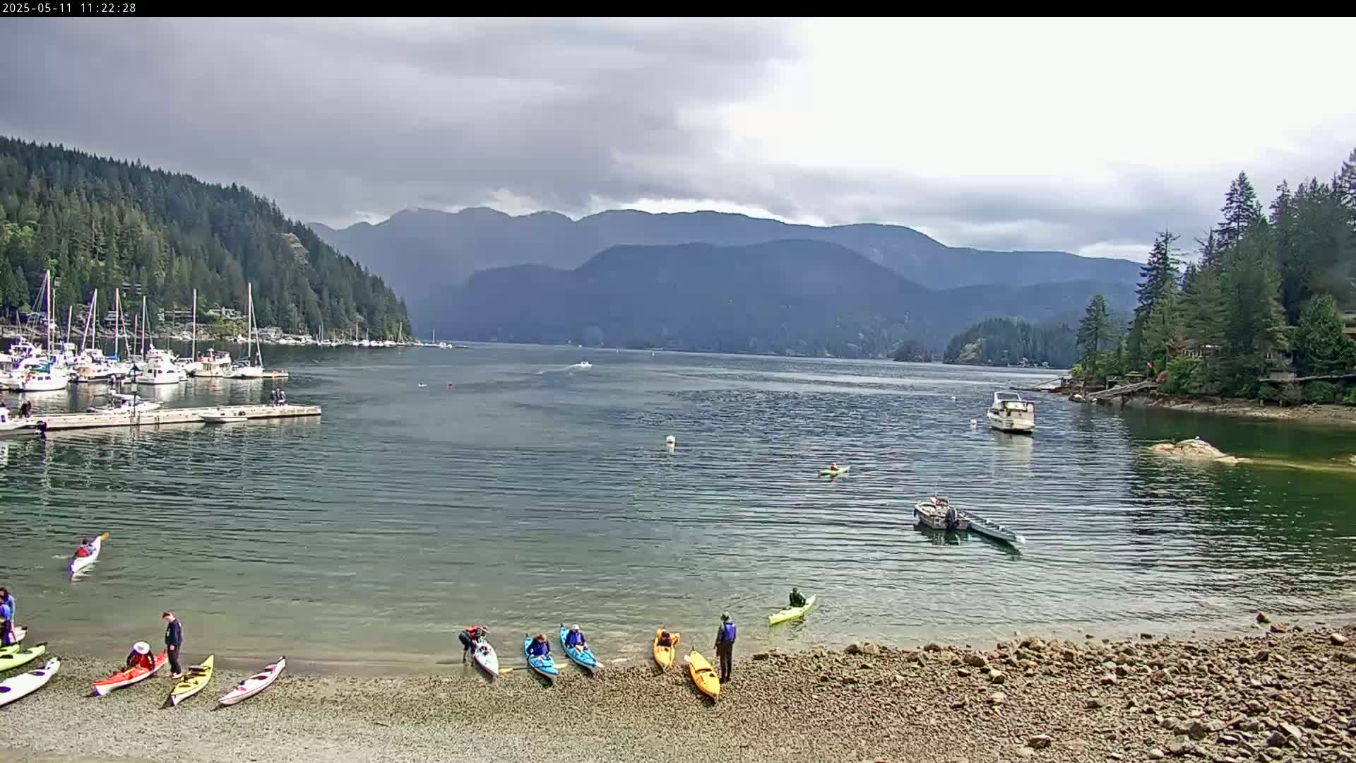 Kayakers are on a rocky beach near a calm bay with boats docked and mountains in the background under a cloudy sky.