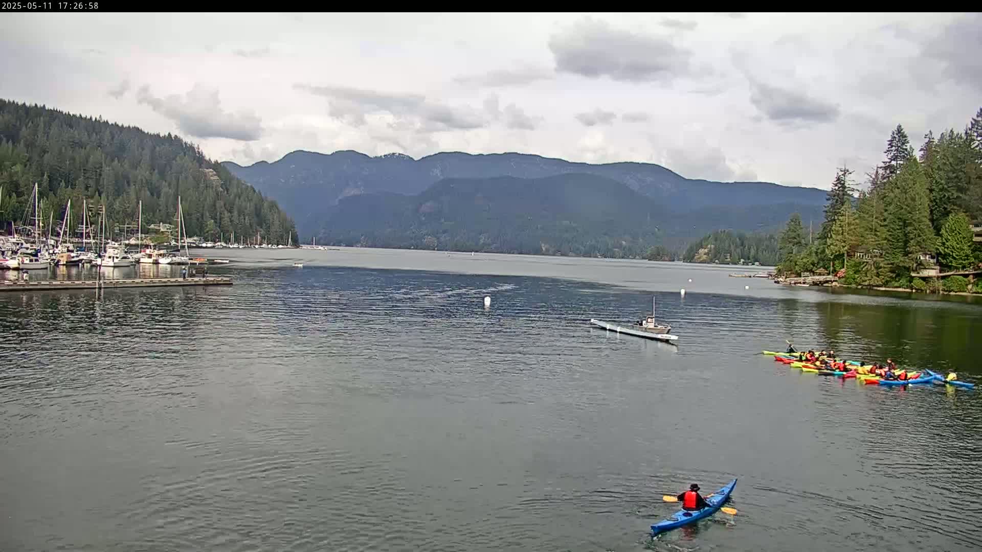 A calm body of water, under a partly cloudy sky, shows several boats docked at a marina near a forested shoreline, with a group of kayakers and a single kayaker paddling in the water against a mountainous backdrop.