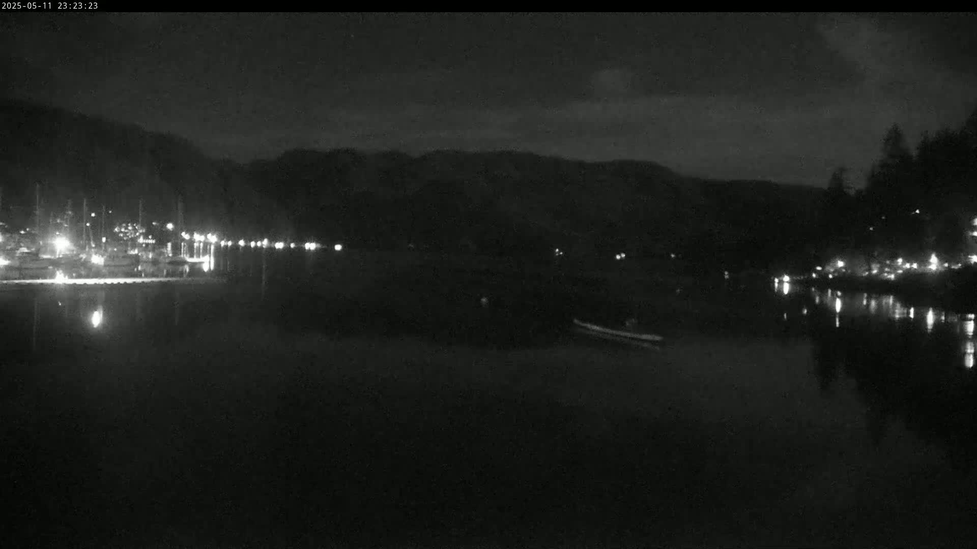 A nighttime view of a calm body of water reflecting the lights of a town nestled at the base of dark hills under a mostly clear sky.
