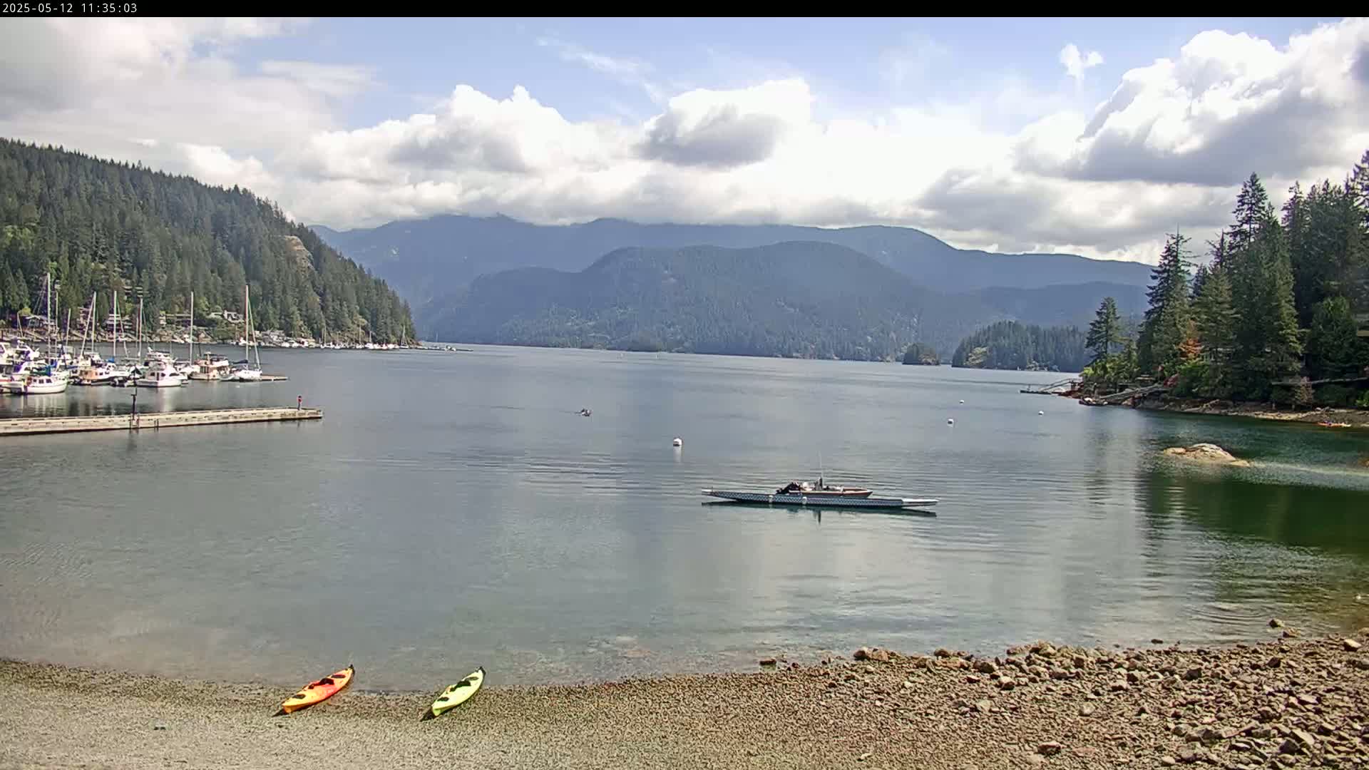 A mostly sunny day shows a calm bay with a rocky shore, two kayaks on the beach, a long boat on the water, and numerous sailboats docked in the distance, all set against a backdrop of forested hills and mountains.