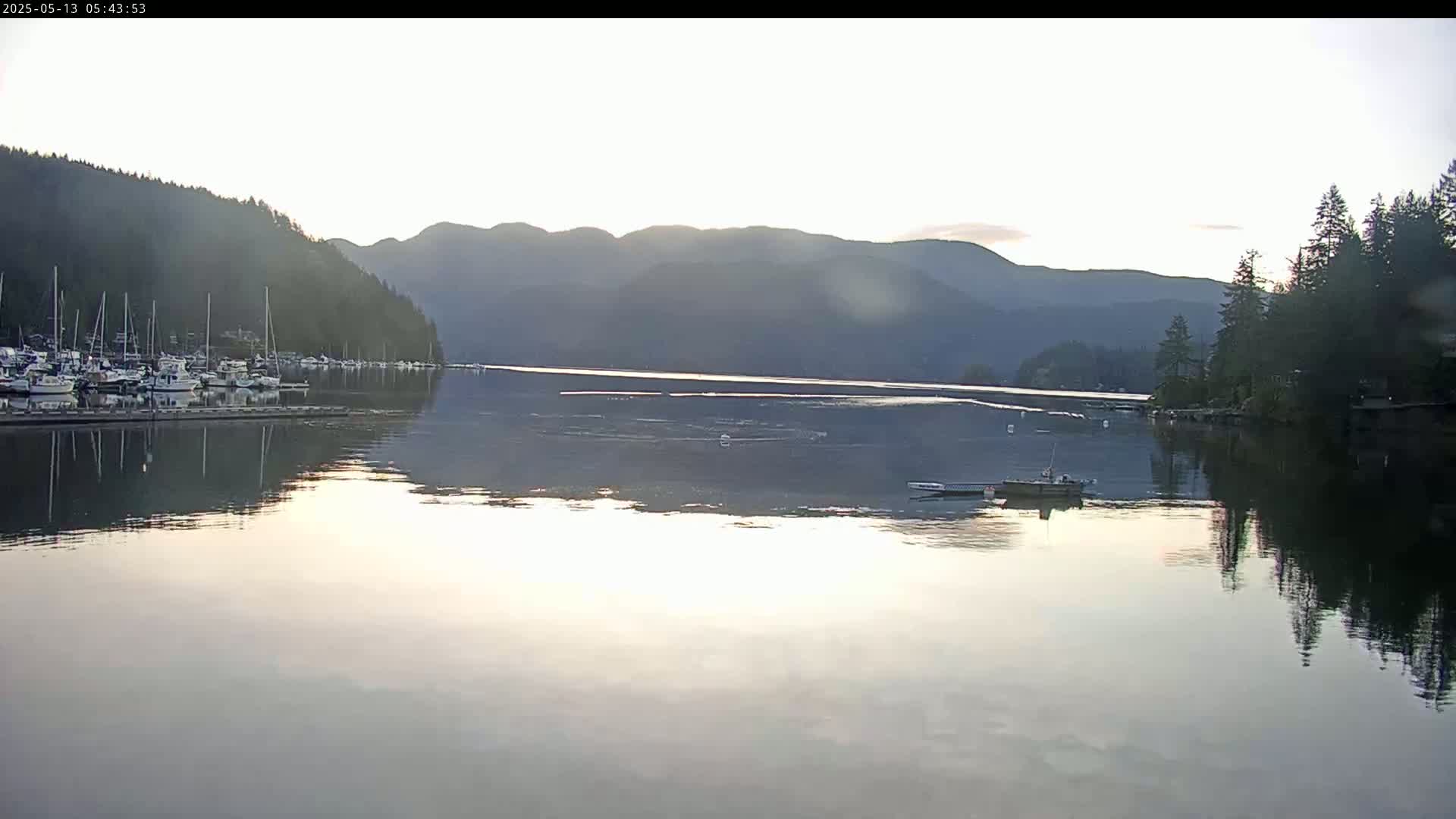 A calm body of water reflects a hazy sunrise and mountains, with several boats docked on one side and a single boat in the middle.