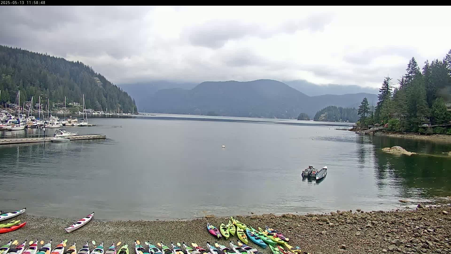 A calm bay under a cloudy sky, with a marina full of boats on one side and a rocky shore lined with kayaks on the other, backed by lush green mountains.