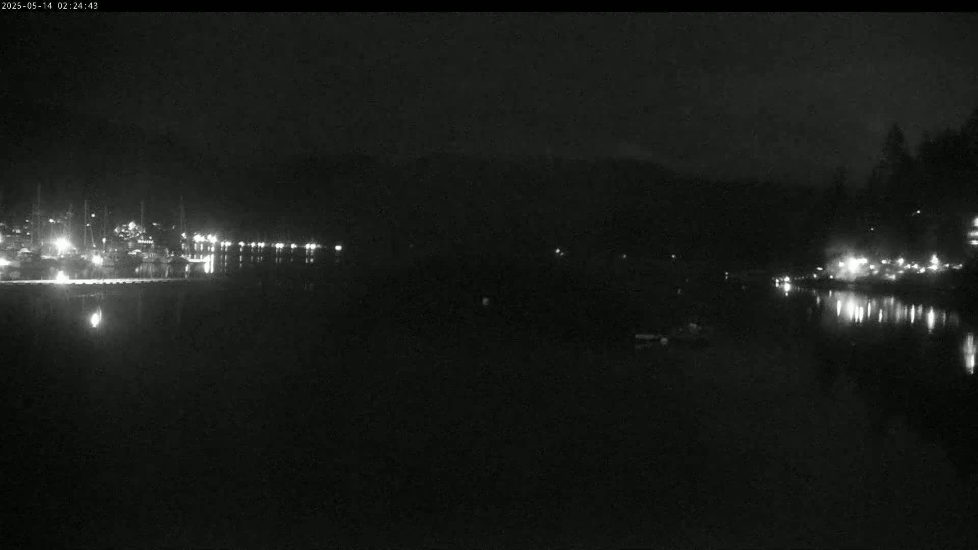 At night, a dark body of water reflects the lights from a distant shoreline with buildings and trees.