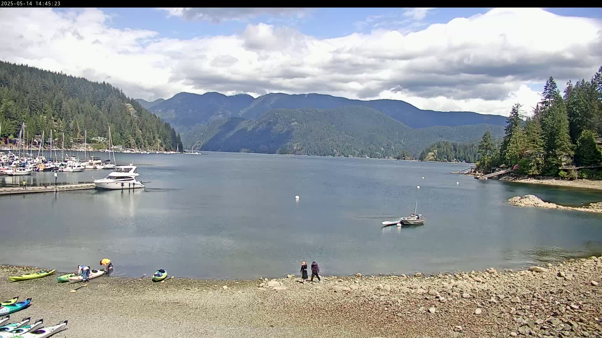 A calm bay with boats and kayaks is nestled between a rocky shoreline and a mountainous background under a partly cloudy sky.