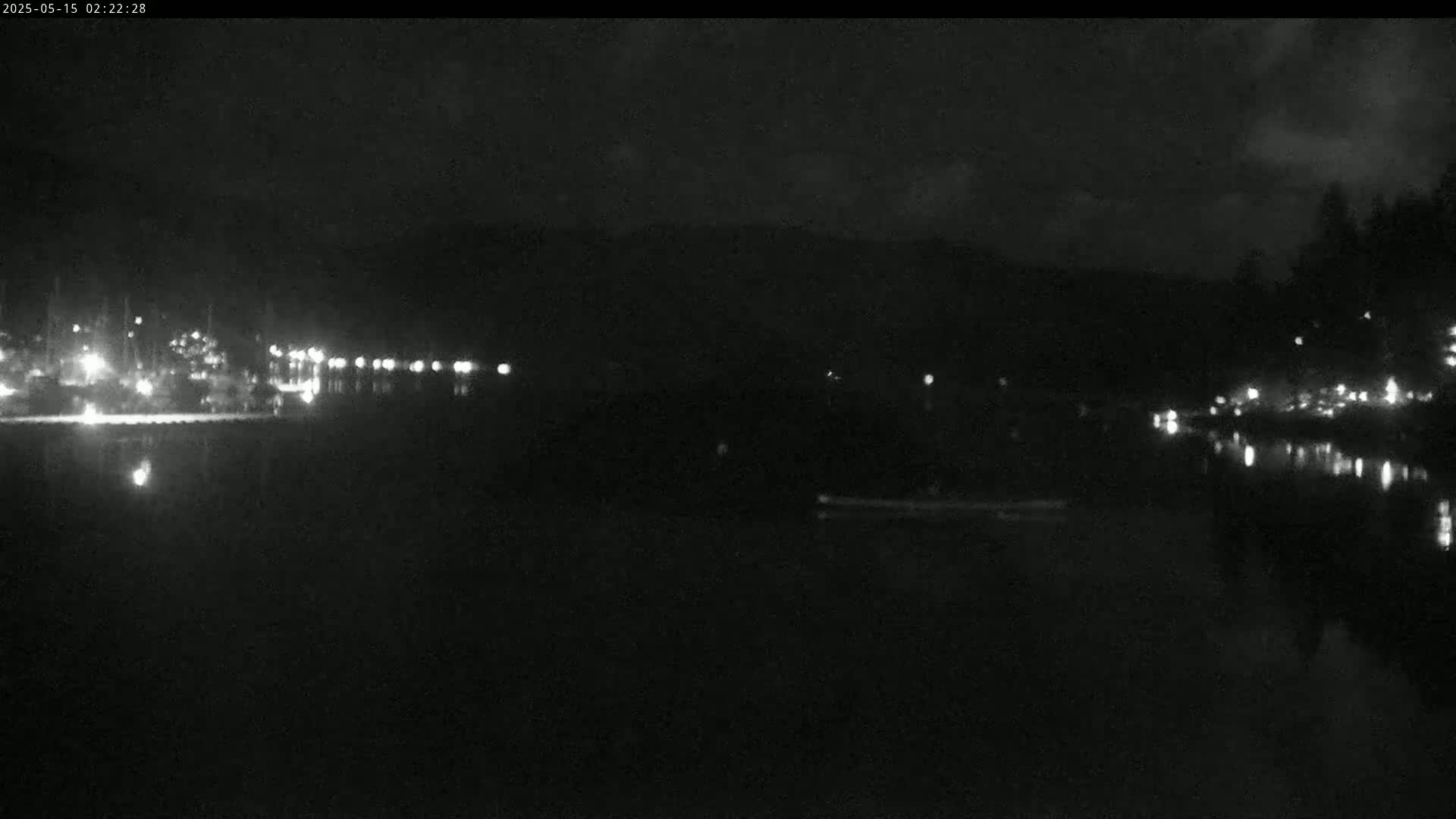 A nighttime, low-light view of a dark body of water with faint lights reflecting on its surface, and a shoreline with sparse lights in the distance.