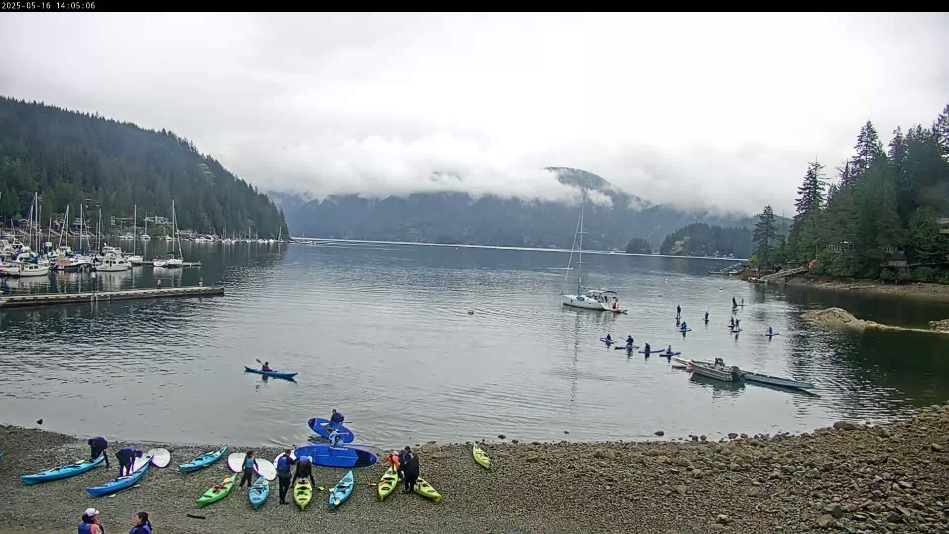 Kayakers and paddleboarders are enjoying a calm, cloudy day on a bay surrounded by forested hills and a marina filled with boats.