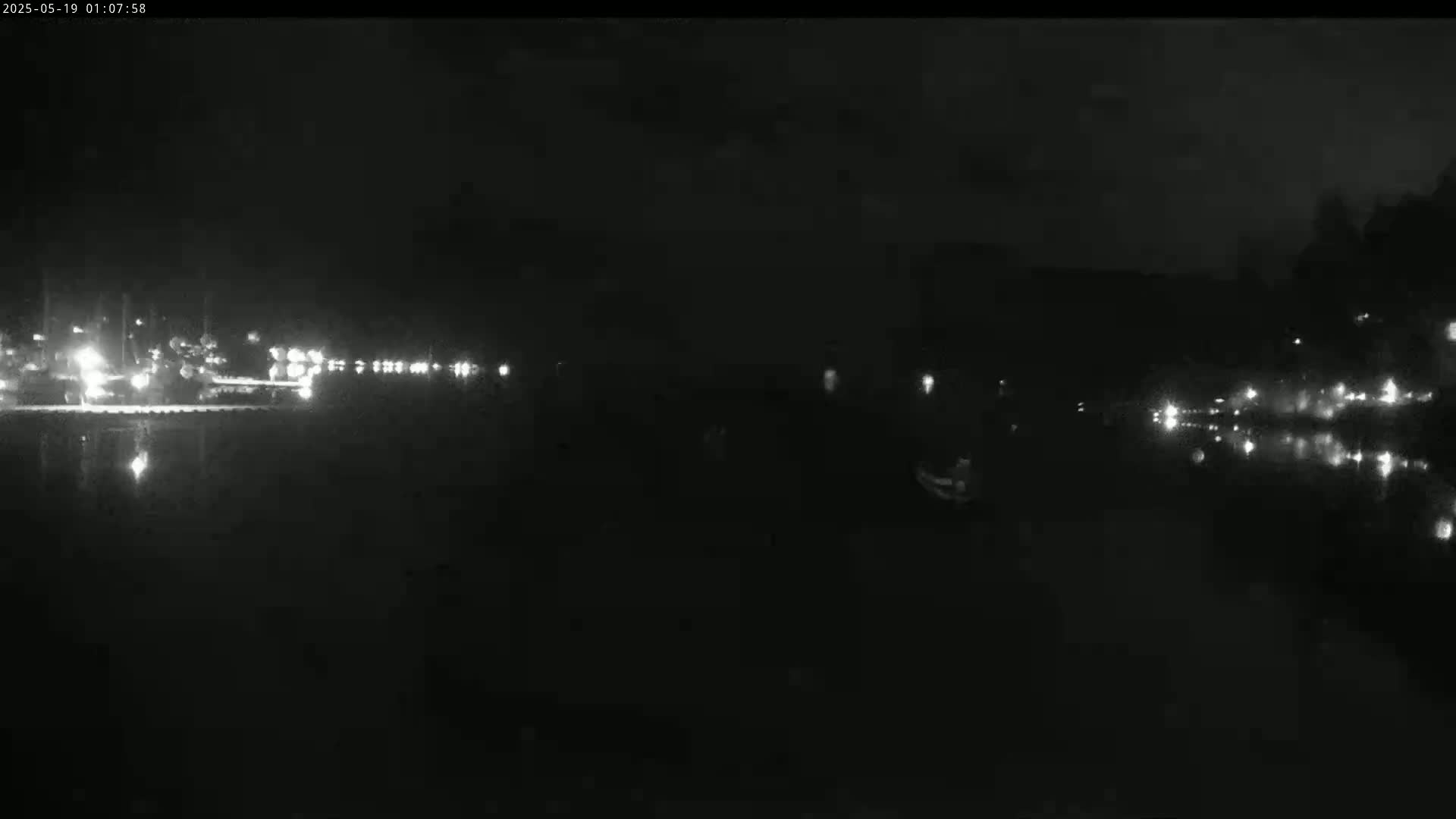 A nighttime, low-light image shows a dark body of water reflecting the lights of vehicles and buildings in the distance.