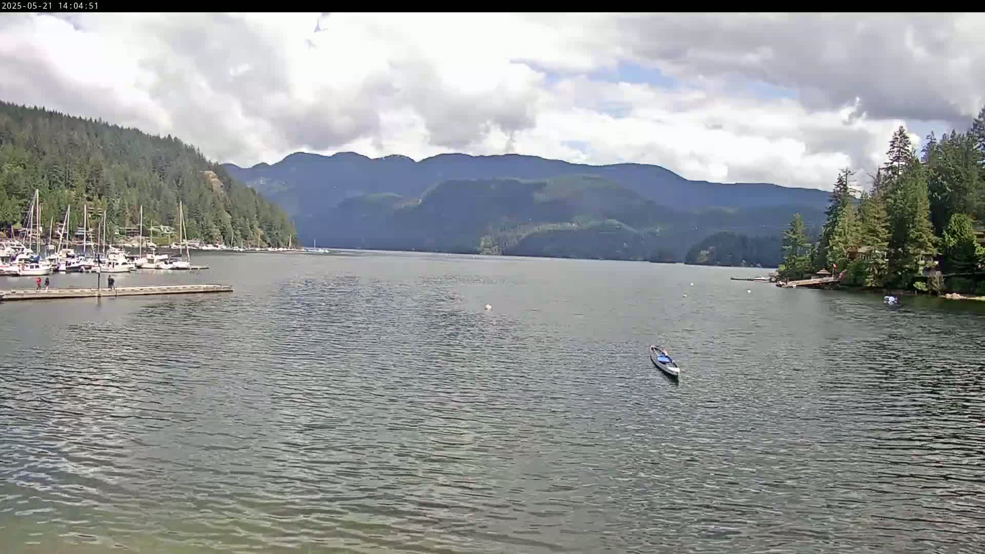 A calm lake with a small boat and a marina is nestled between tree-covered hills under a partly cloudy sky.