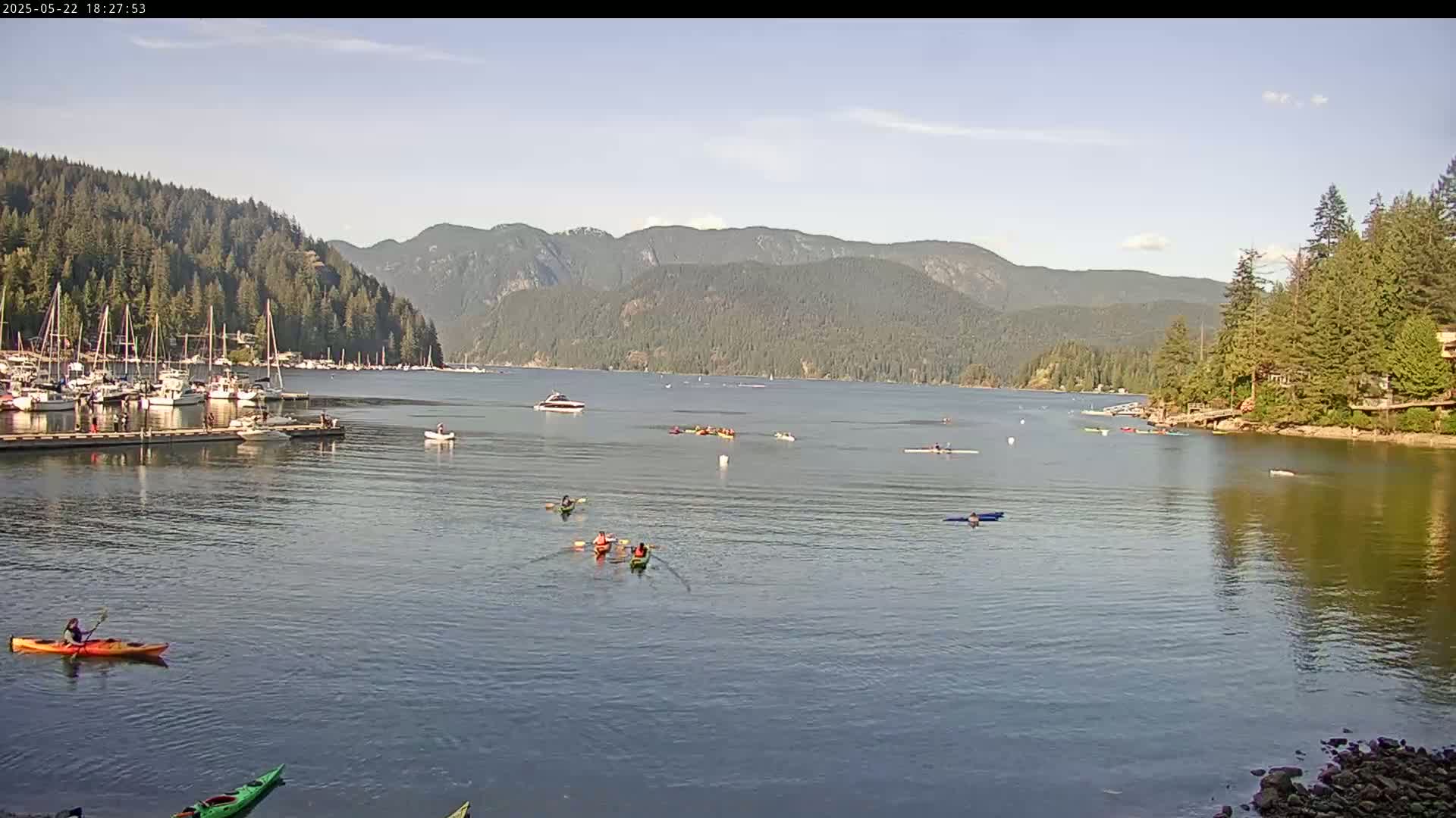 Kayakers paddle on a calm bay surrounded by forested hills under a mostly sunny sky, with sailboats moored at a dock in the foreground.