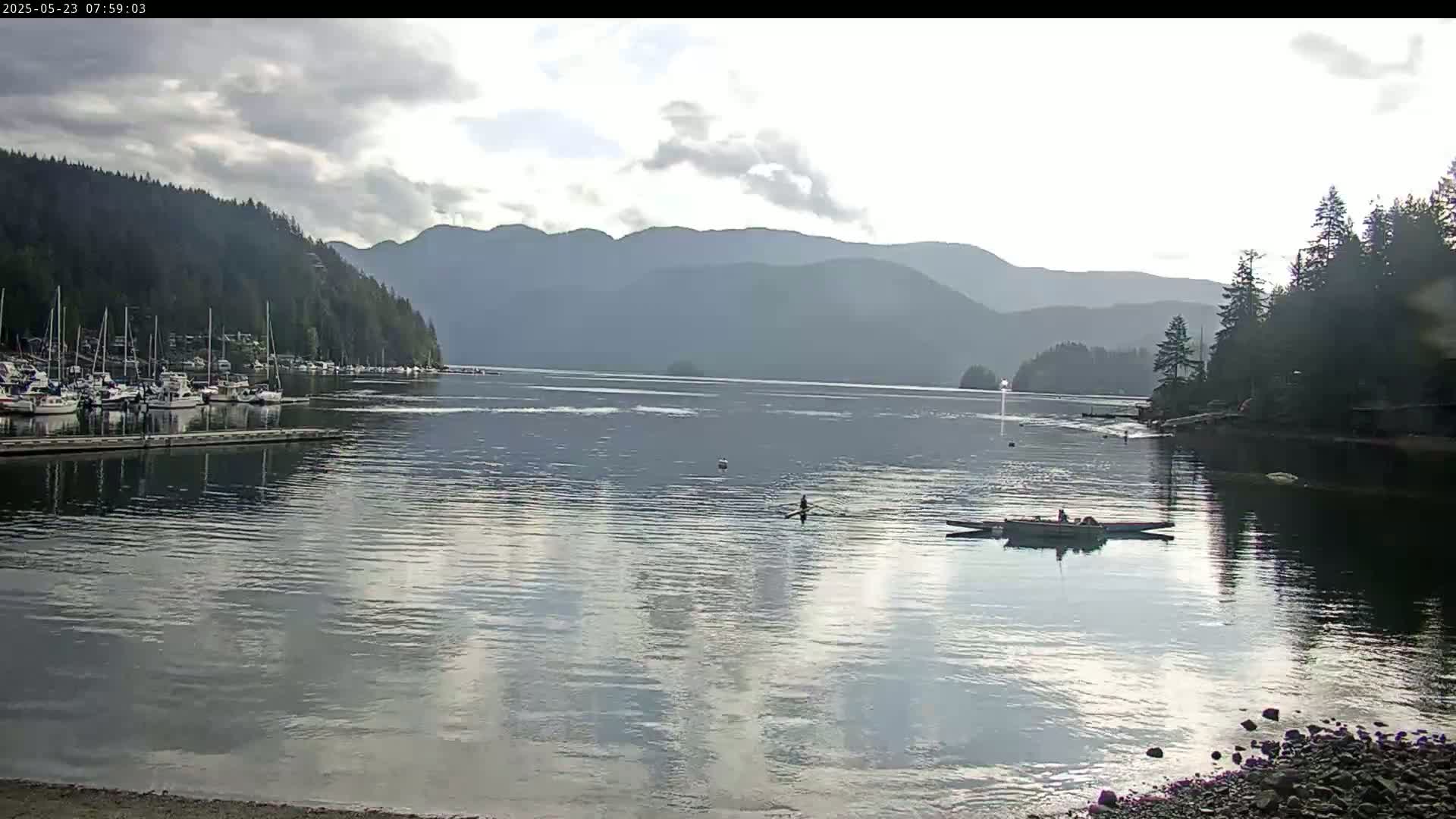 A calm bay with a marina full of boats, a few people in a rowboat, and mountains in the background under a partly cloudy sky.