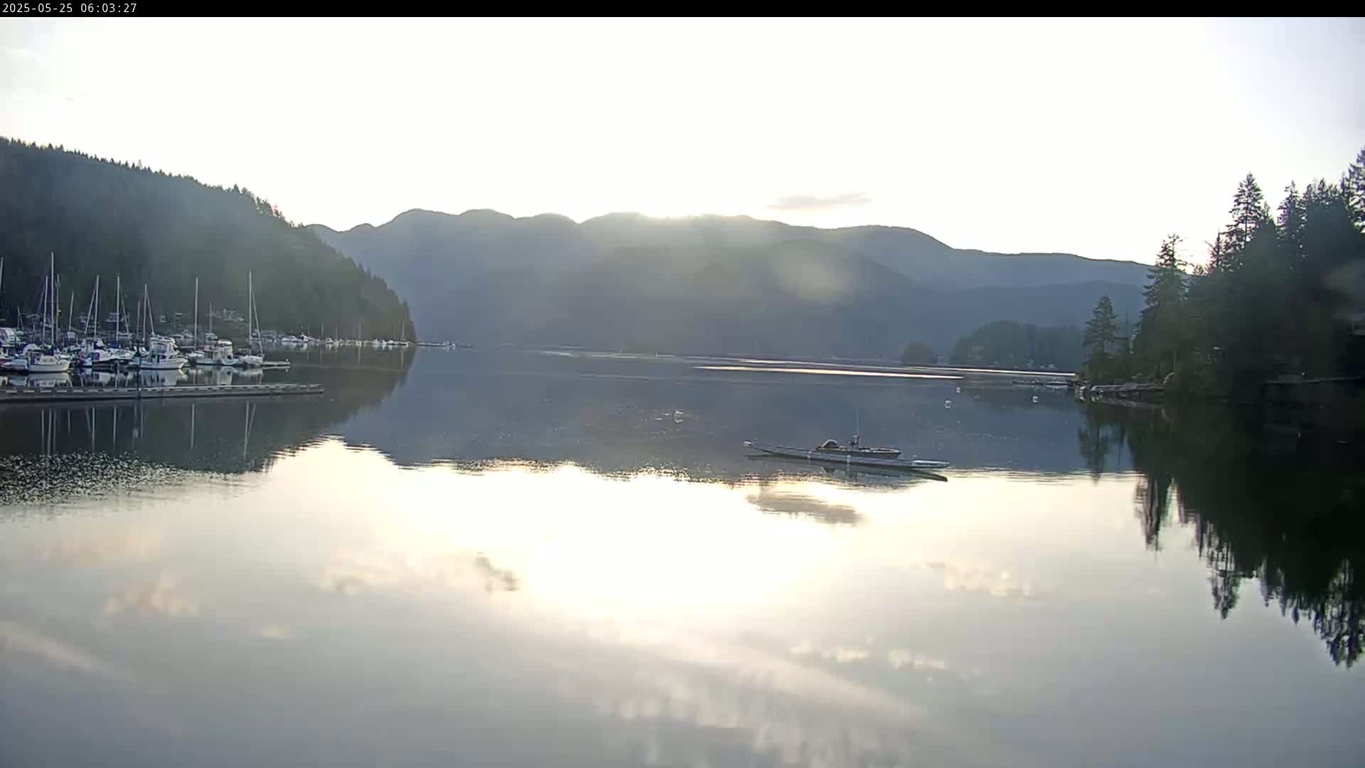 Calm water reflects a sunrise over a marina nestled between forested hills.