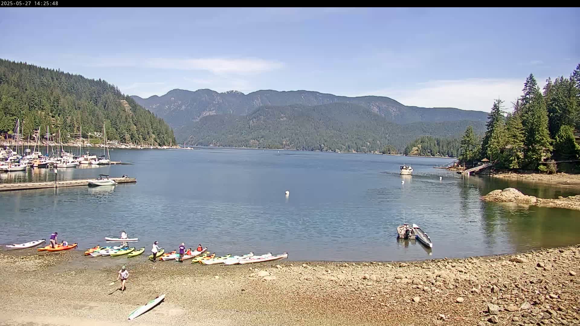A calm, sunny day shows a bay with a rocky shore, numerous kayaks pulled up on the beach, several boats moored in the water, and forested mountains in the background.