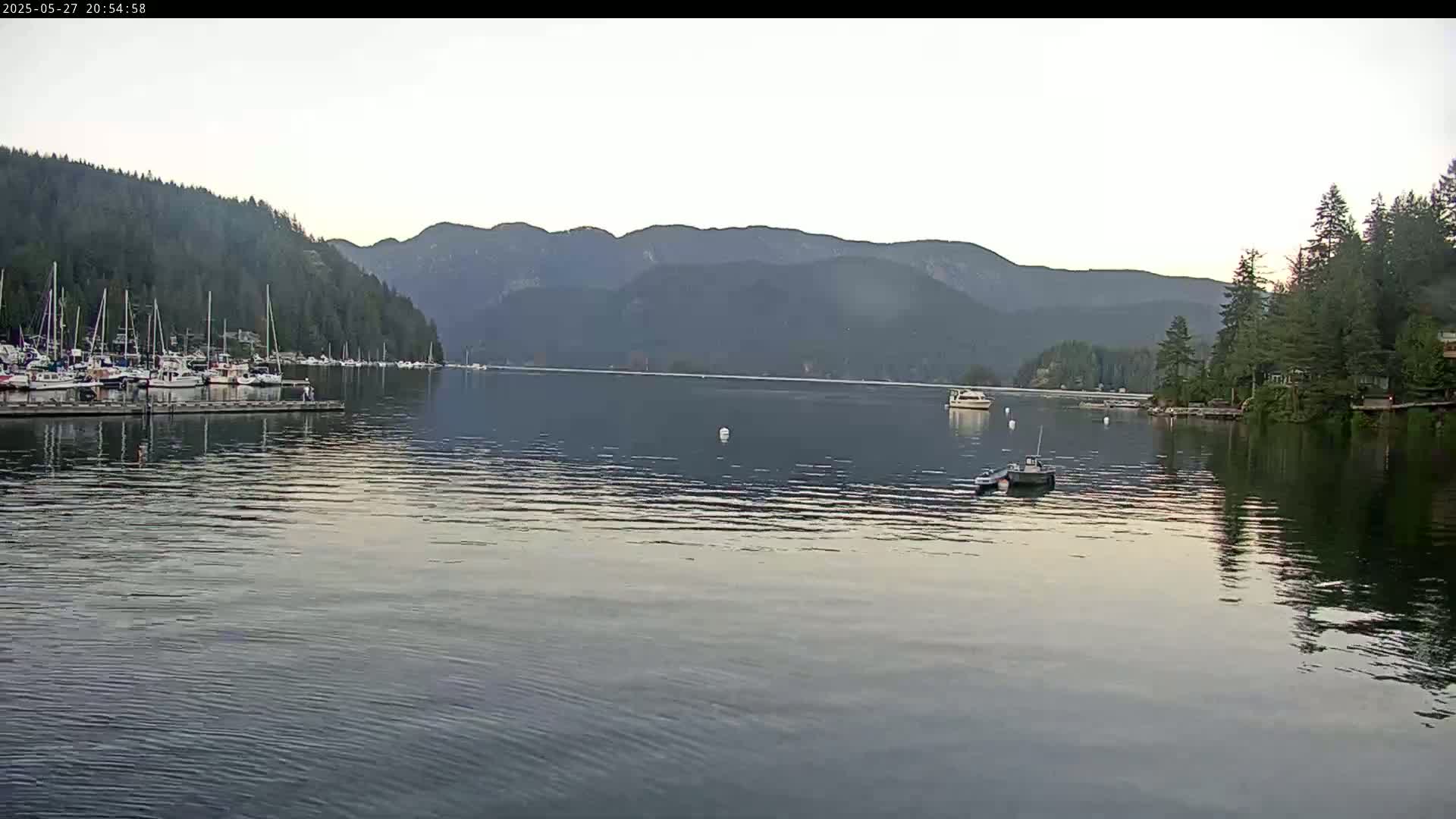 A calm body of water shows a marina with several boats, a few other boats on the water, and forested mountains in the background under a mostly clear sky.