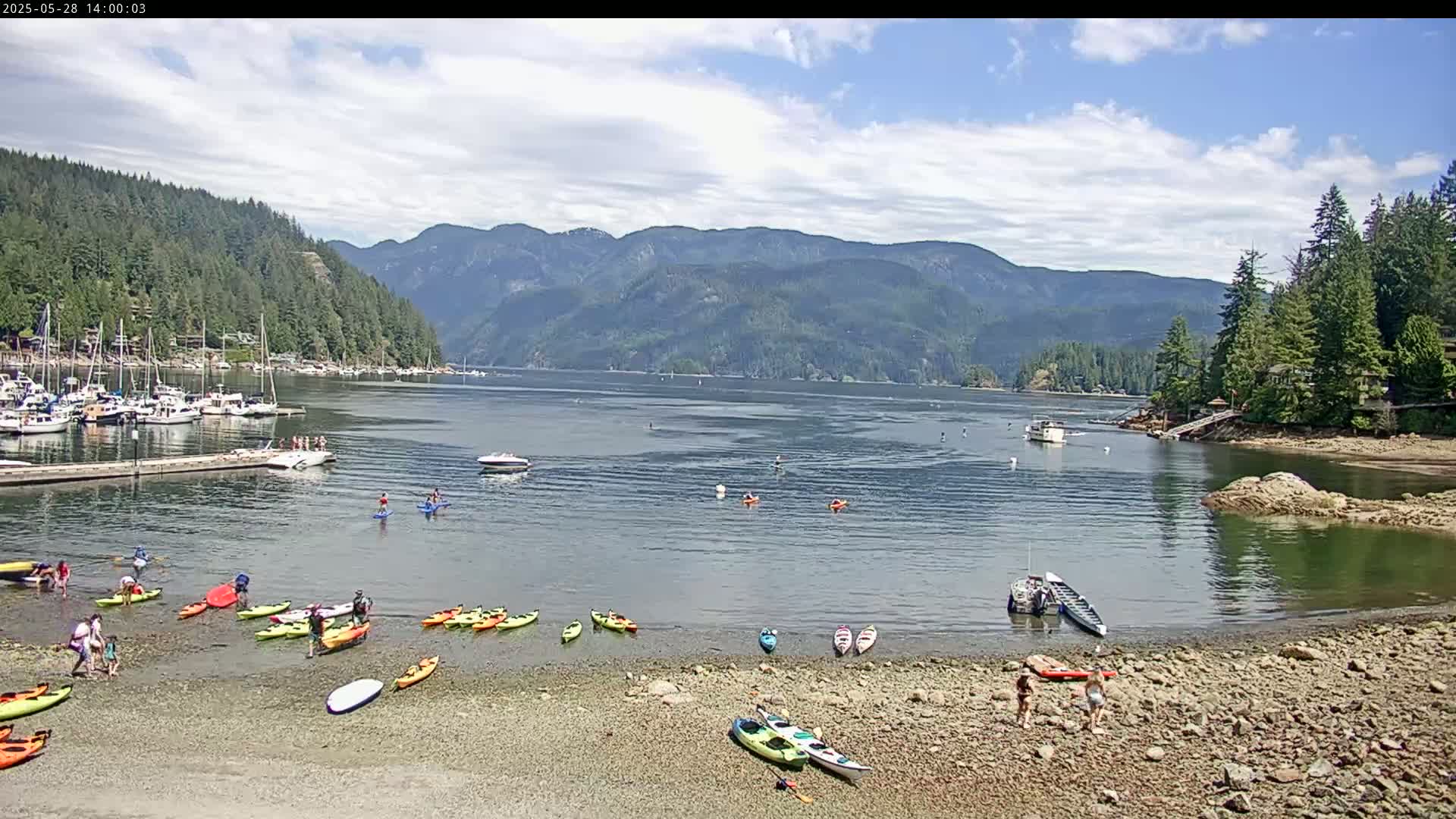 A rocky shoreline with numerous kayaks and people, a calm bay with boats and people in the water, and mountains in the background under a partly cloudy sky.