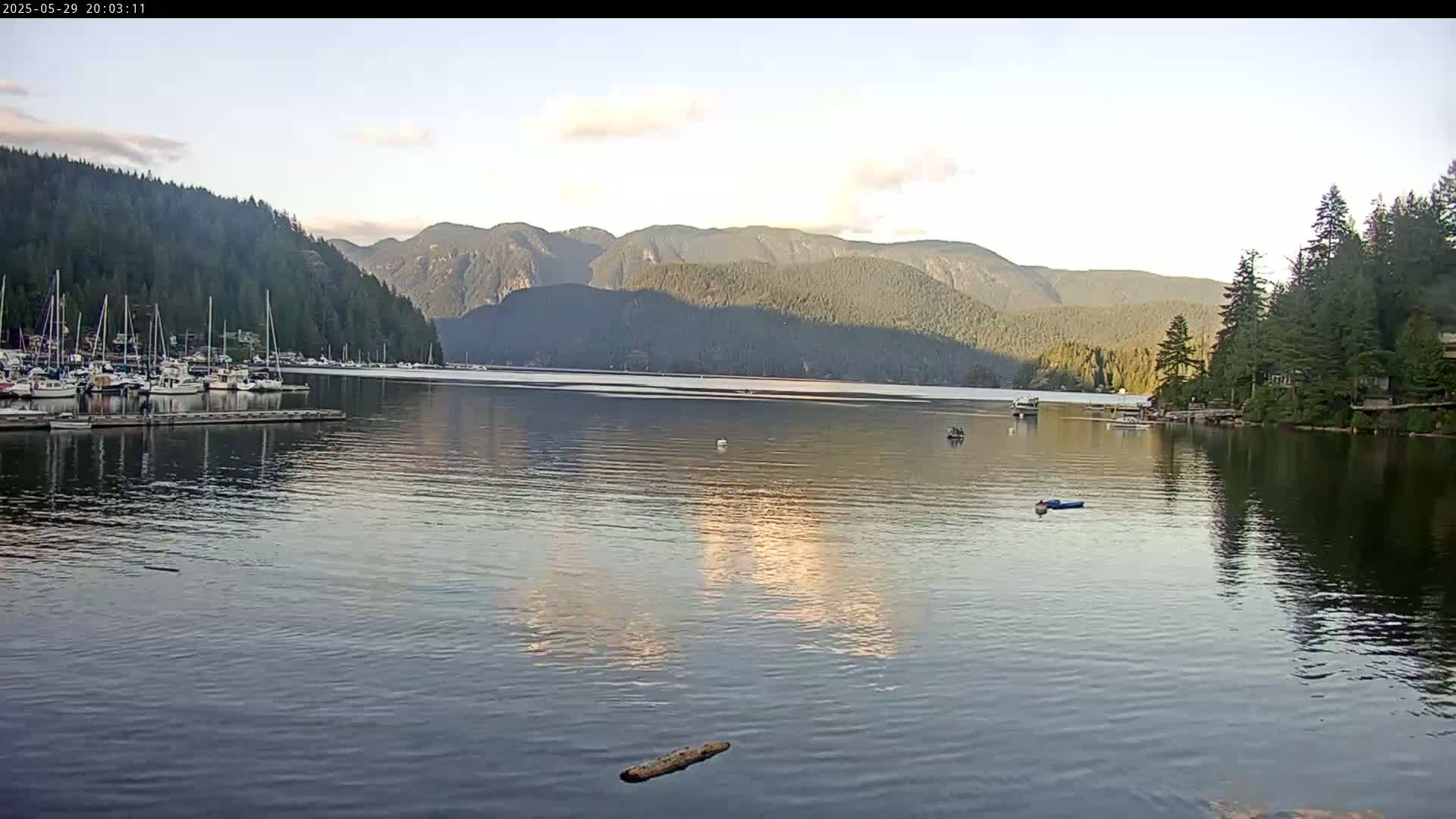 A calm body of water reflects a partly cloudy sky and forested mountains, with a marina full of sailboats to the left.