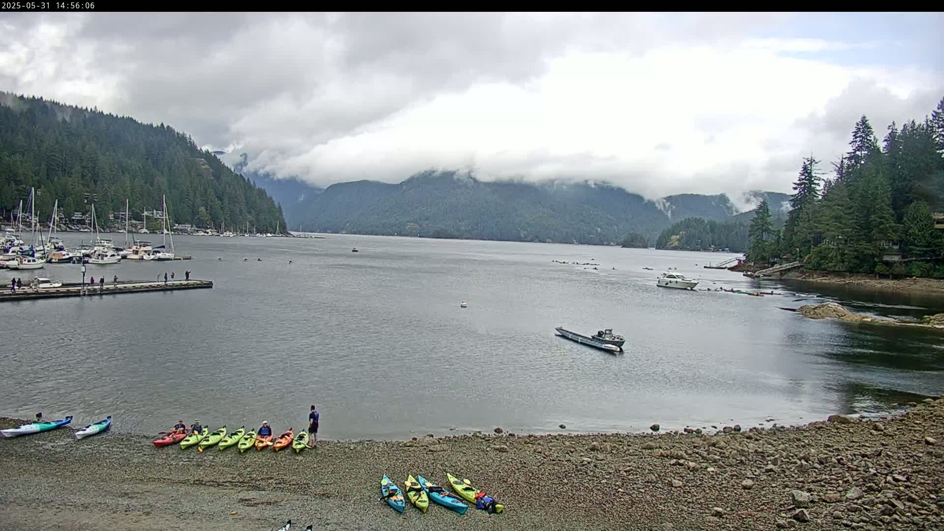 A calm bay under a cloudy sky is surrounded by lush green mountains, with several boats and kayaks on the water and shore.