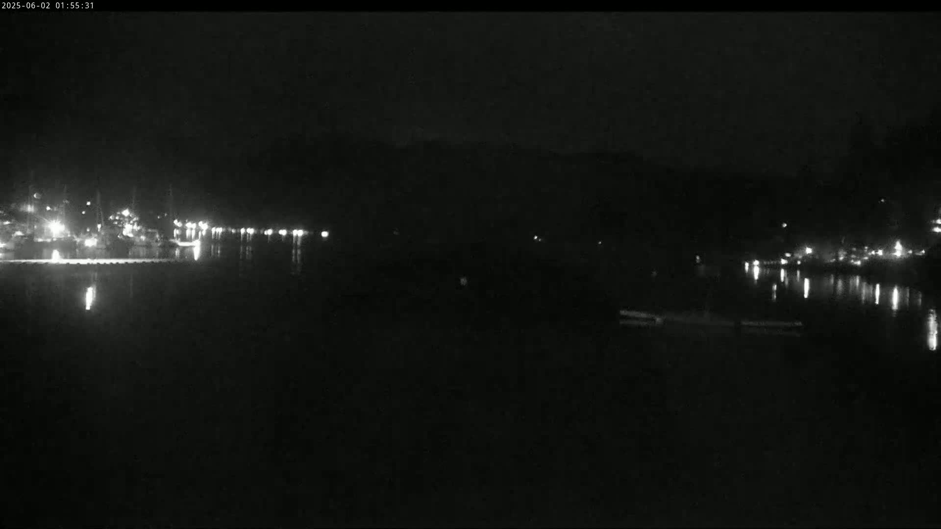 A dark nighttime scene shows a calm body of water reflecting the faint lights of a distant shoreline.