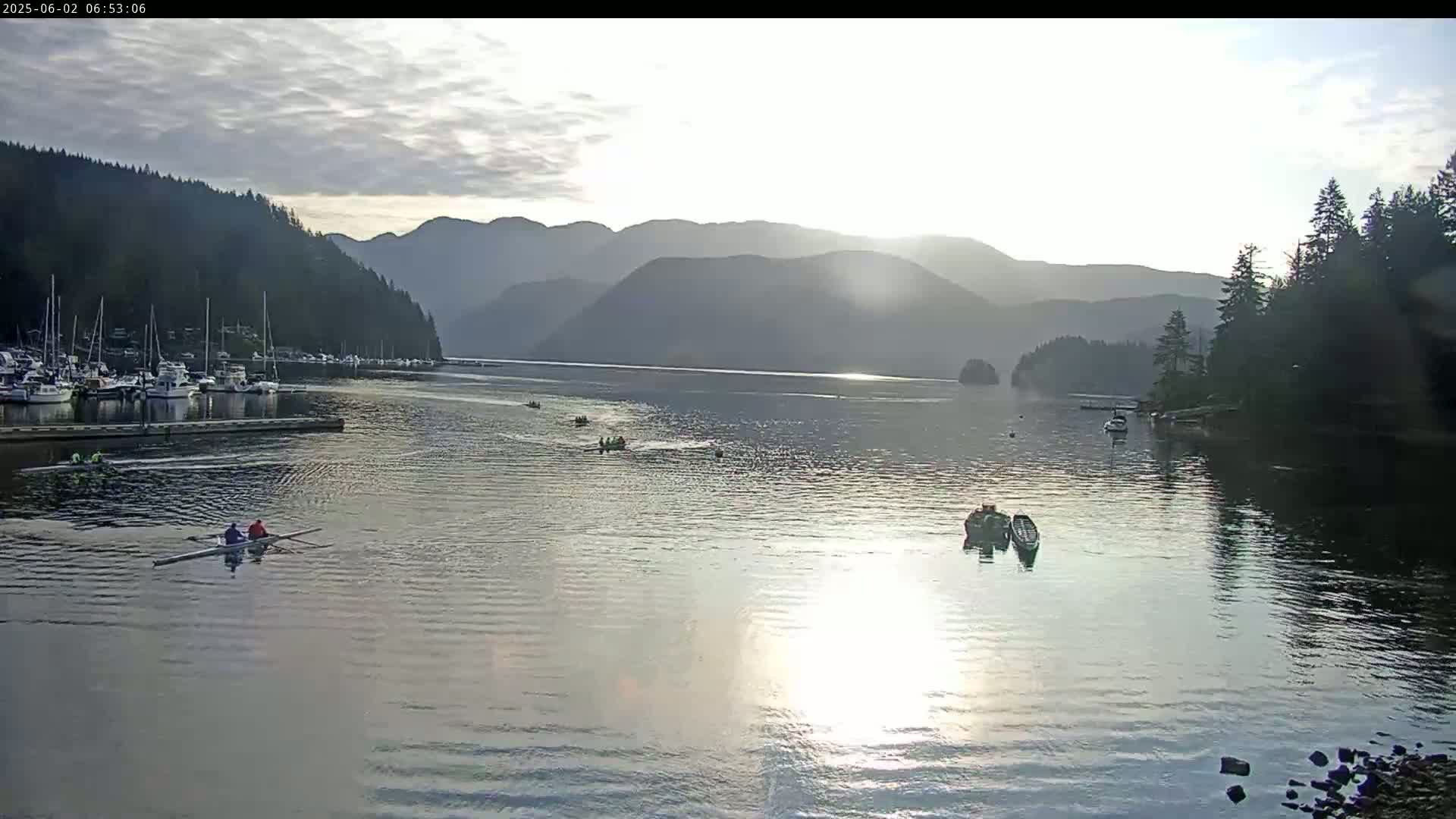 Several rowing sculls are on a calm, sunlit bay surrounded by forested mountains under a mostly cloudy sky.