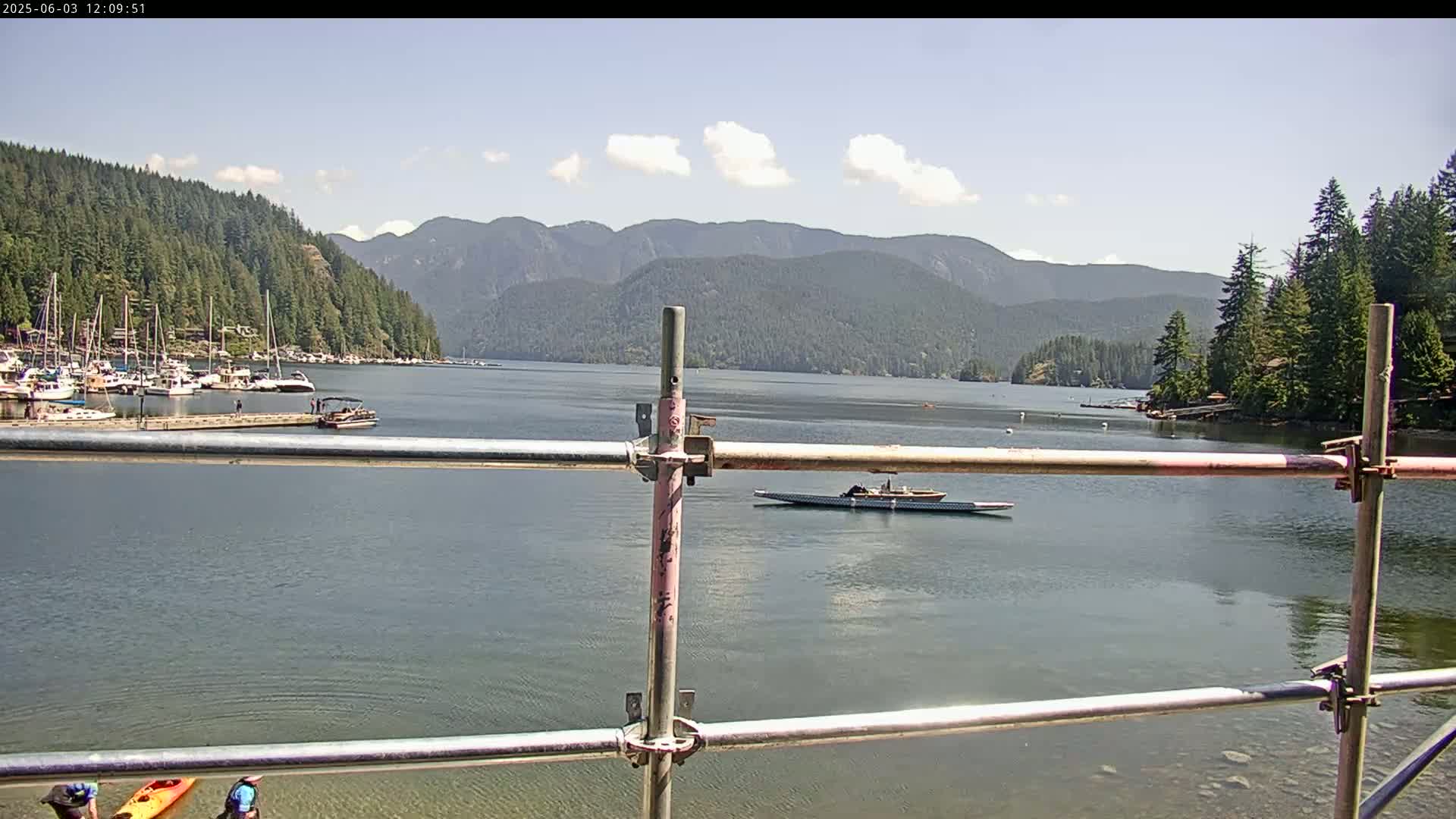 A calm body of water with a marina, a rowing shell, and kayakers, situated in front of a mountainous landscape under a partly cloudy sky.