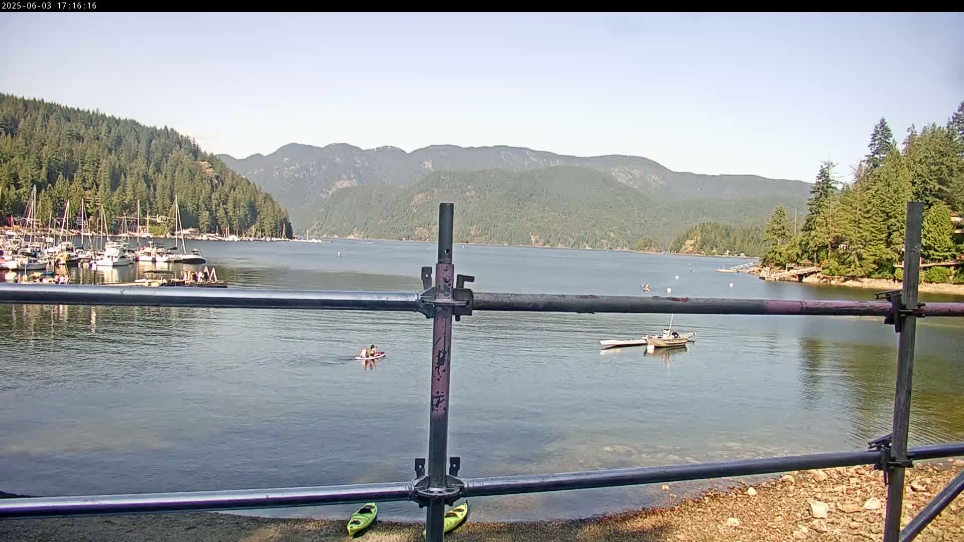 A calm bay with several boats and people swimming or kayaking, viewed from behind a metal railing, is set against a backdrop of forested mountains under a clear sky.