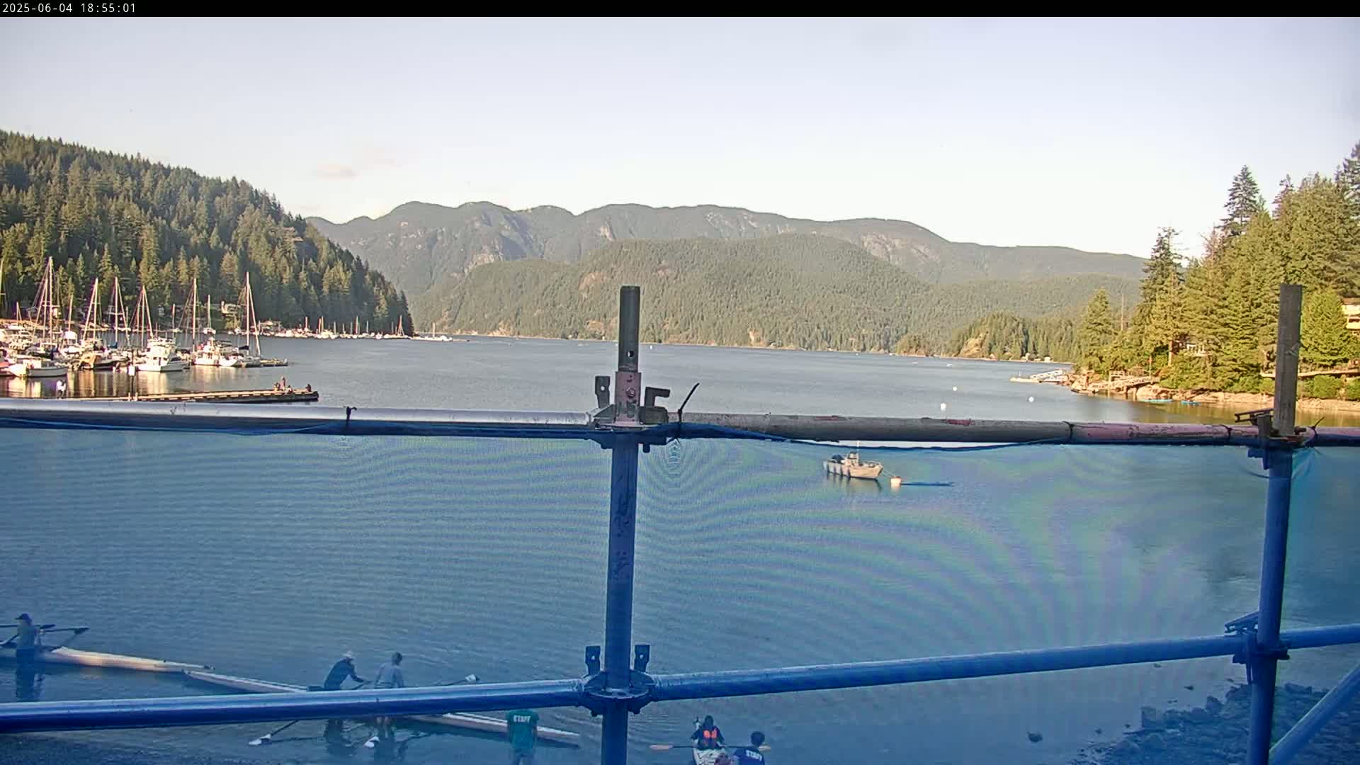 A calm bay with numerous sailboats in a marina, surrounded by lush green mountains under a clear sky, is partially obscured by a blue construction fence with people near several rowboats in the foreground.