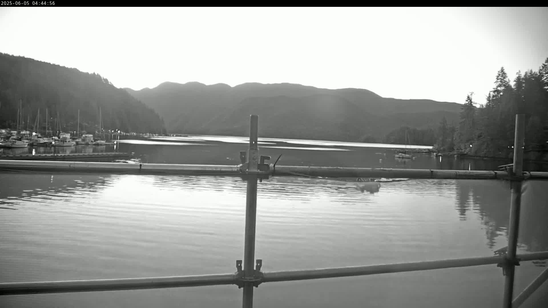 A calm body of water with a marina and mountains in the background under a bright, overcast sky is seen through a metal scaffolding structure.