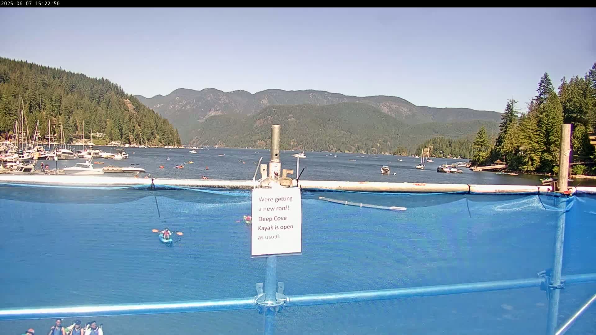 A calm bay with numerous boats and kayakers is visible beyond a blue tarp enclosure on a sunny day, with mountains in the background.