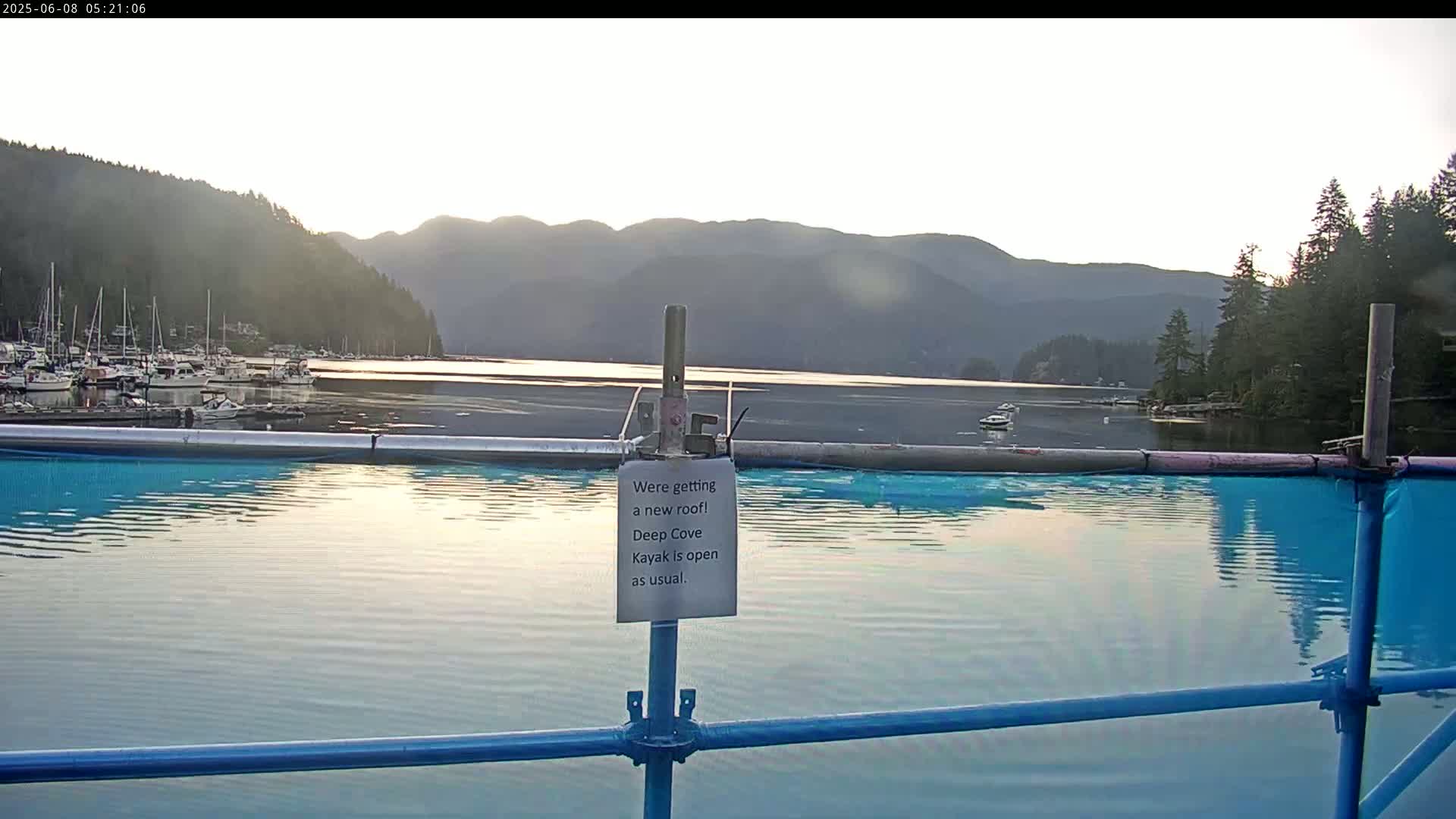 A calm body of water reflects the morning light, showcasing a marina with numerous boats and forested mountains in the background under a bright, clear sky.