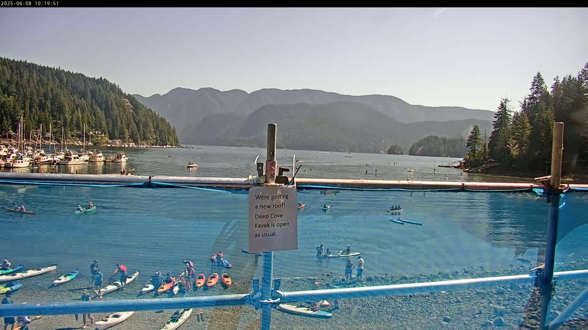 A calm, sunny day shows a cove with numerous kayaks and paddleboards in the water, along with a marina full of boats in the background, all set against a backdrop of mountains.