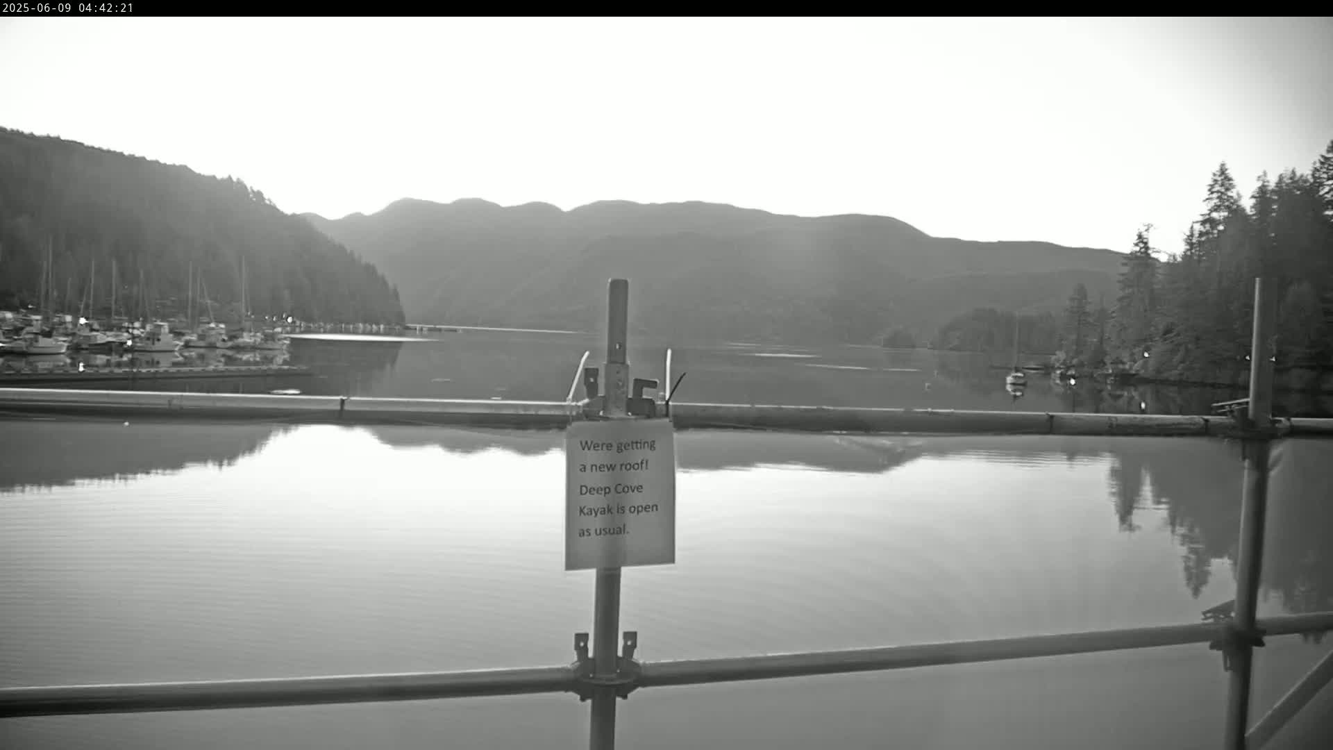 A calm body of water reflects a mountainous landscape and a marina under a bright, overcast sky.