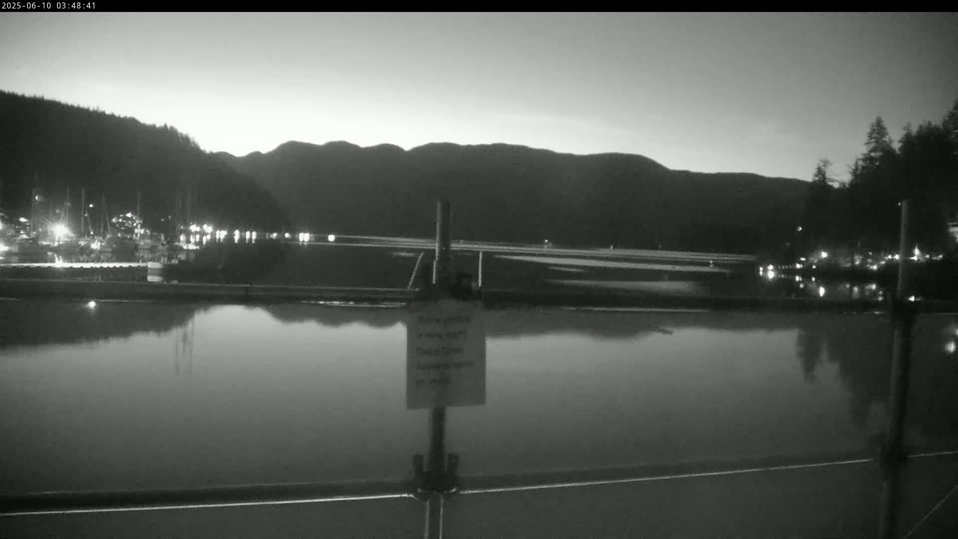 A calm body of water reflects the lights of a harbor at dusk, with dark mountains in the background under a clear sky.