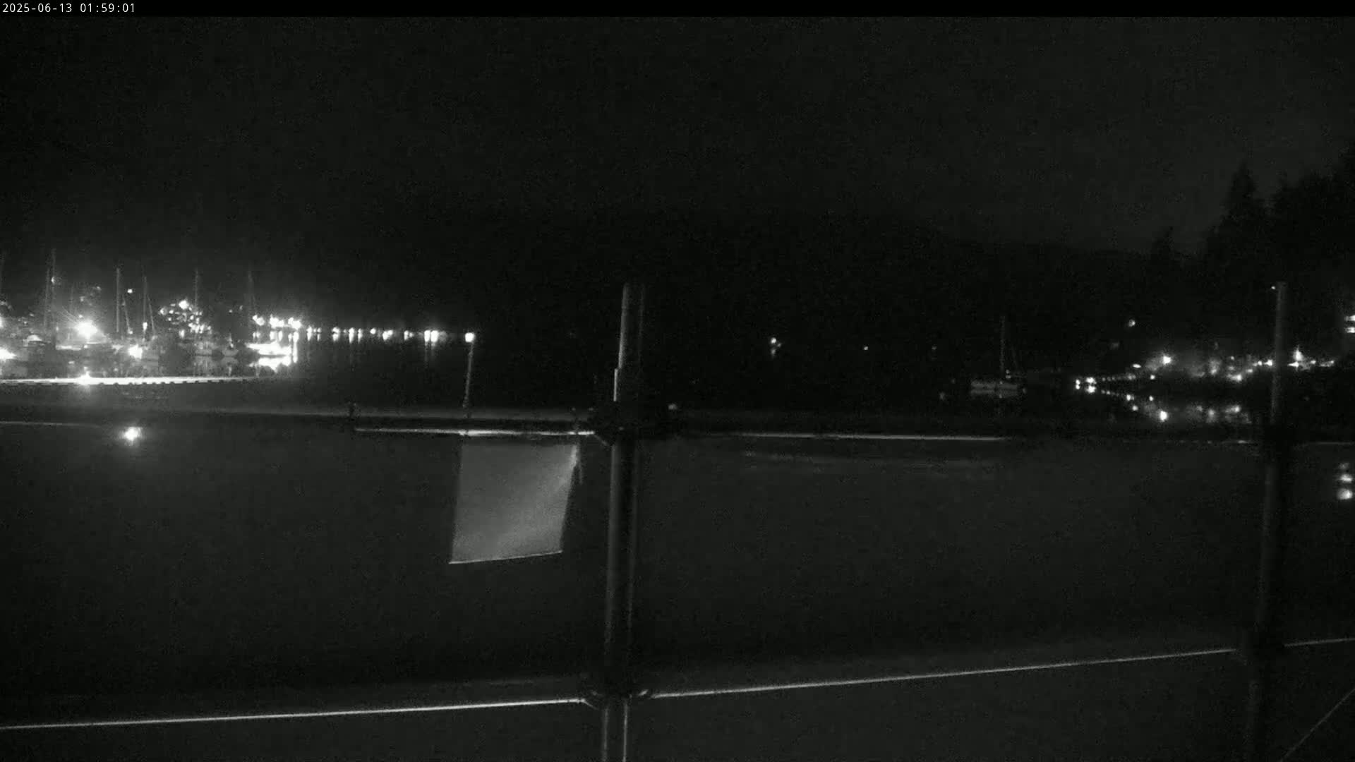 A nighttime view of a calm body of water with boats docked and lights visible across the water, seen from behind a dark fence.