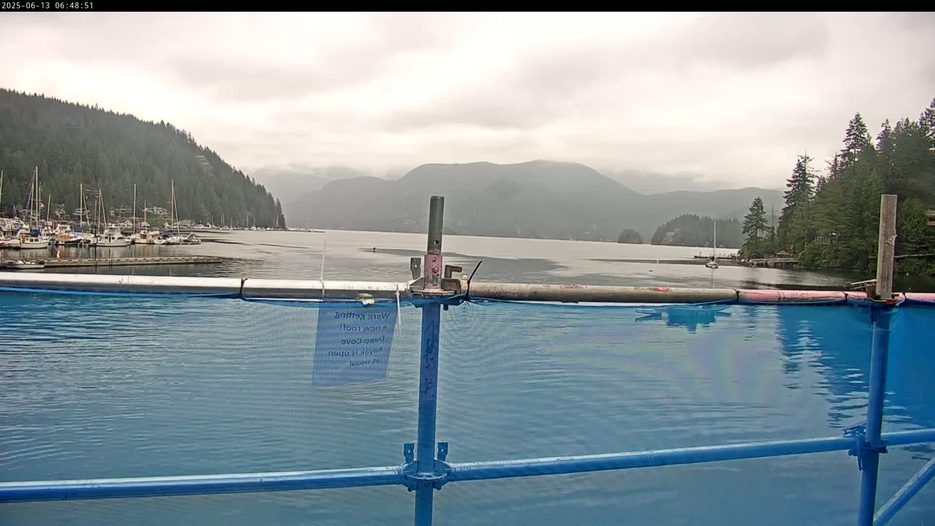 A calm body of water with boats in a marina is partially obscured by a blue construction barrier, all set against a backdrop of overcast mountains and forested hills.