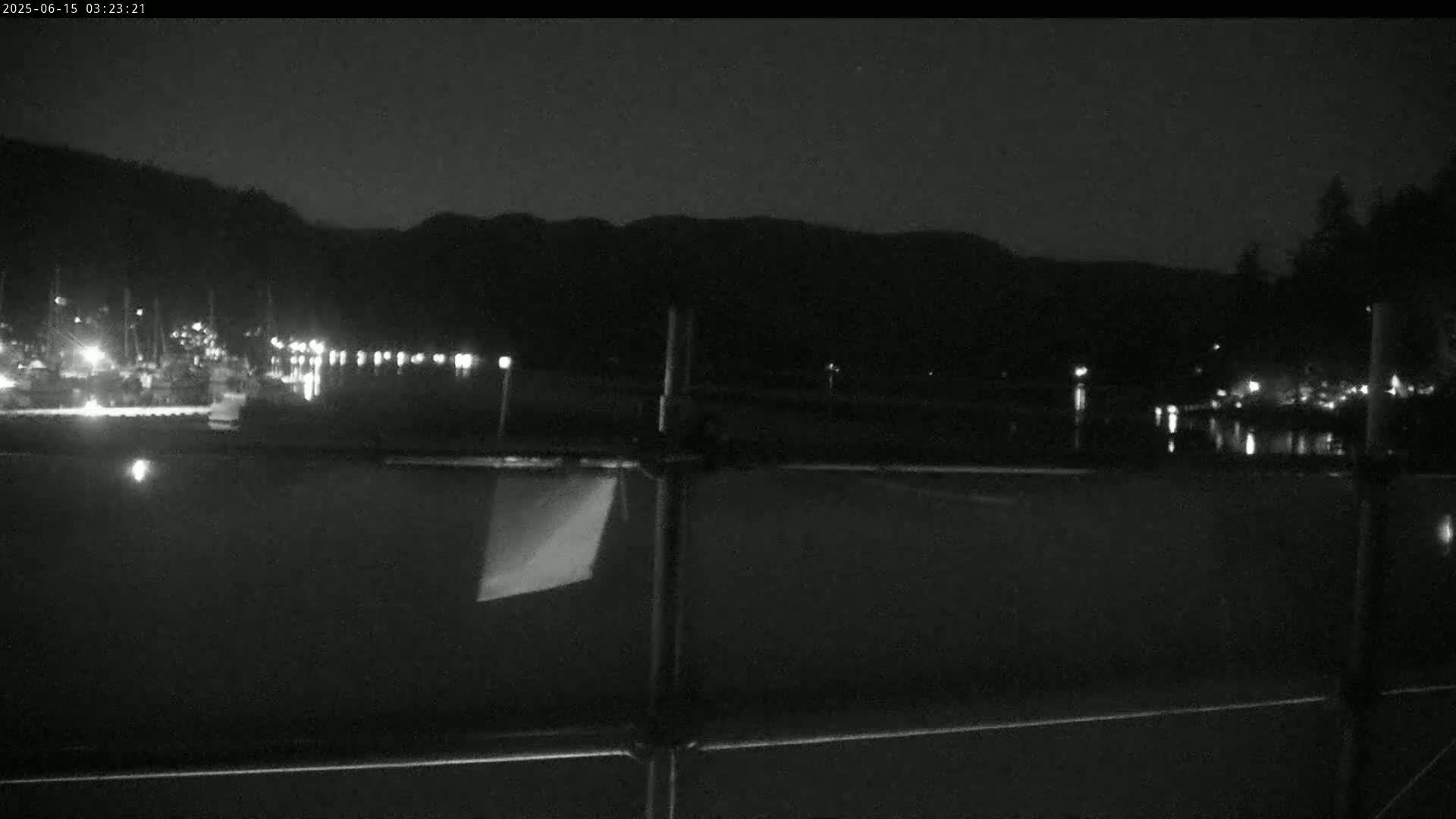 A nighttime view of a calm body of water with boats docked along a shoreline, partially obscured by a metal fence in the foreground, under a dark sky.