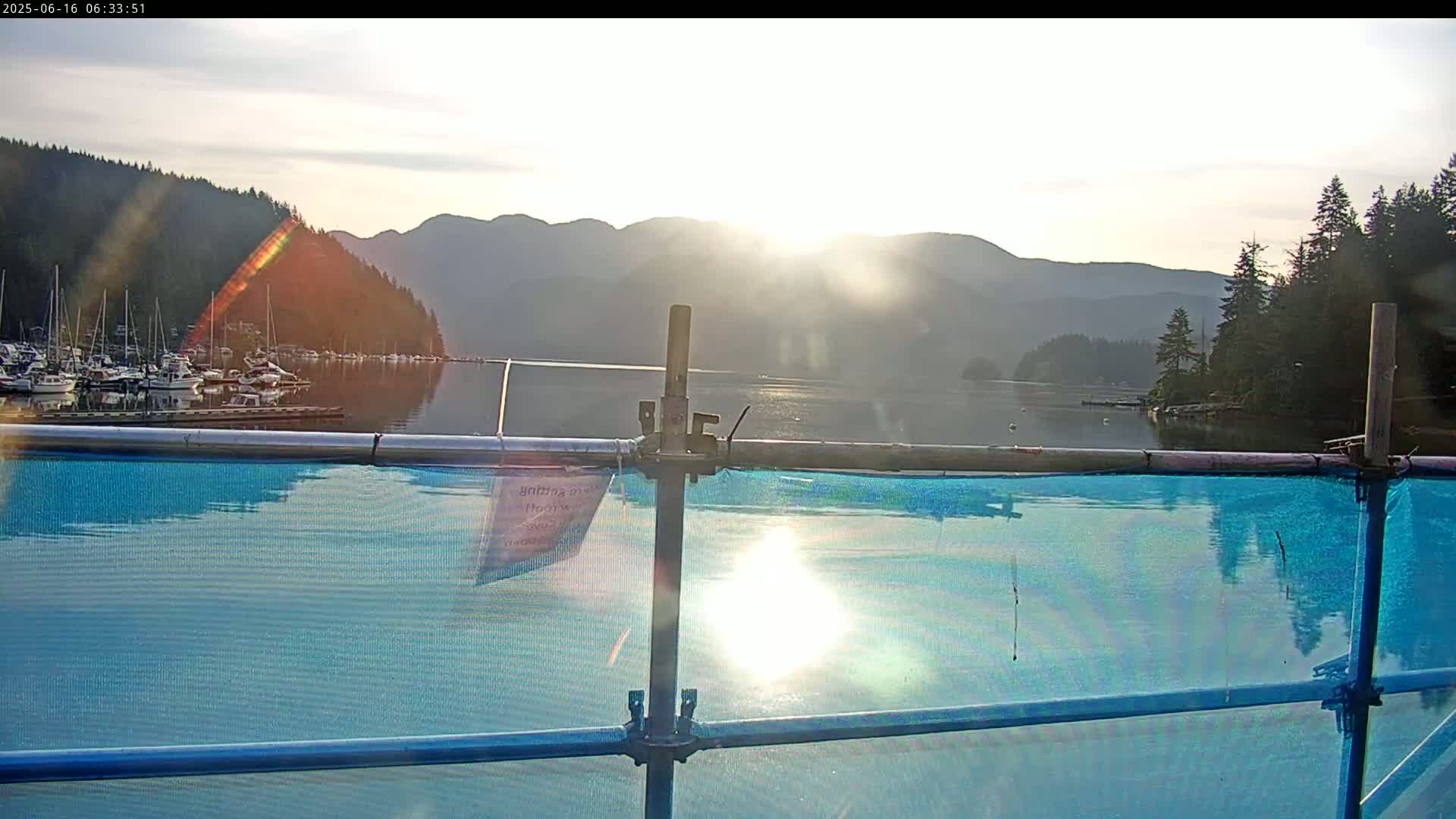 A bright, sunlit scene of a calm body of water with numerous boats docked, mountains in the background, and a blue tarp-covered scaffolding in the foreground.