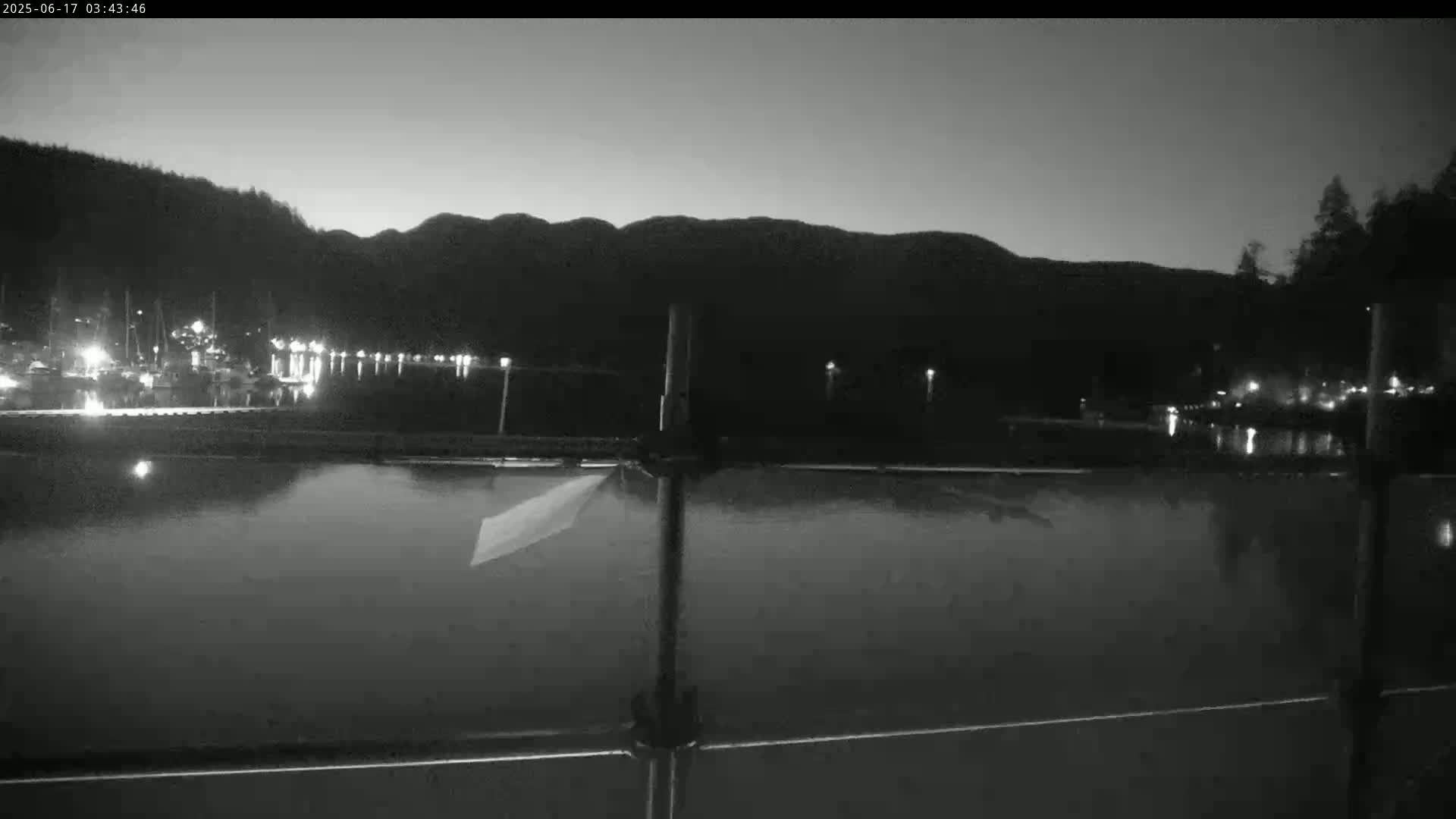 A dark, calm body of water reflects the lights of a marina nestled against a silhouetted mountain range under a twilight sky.