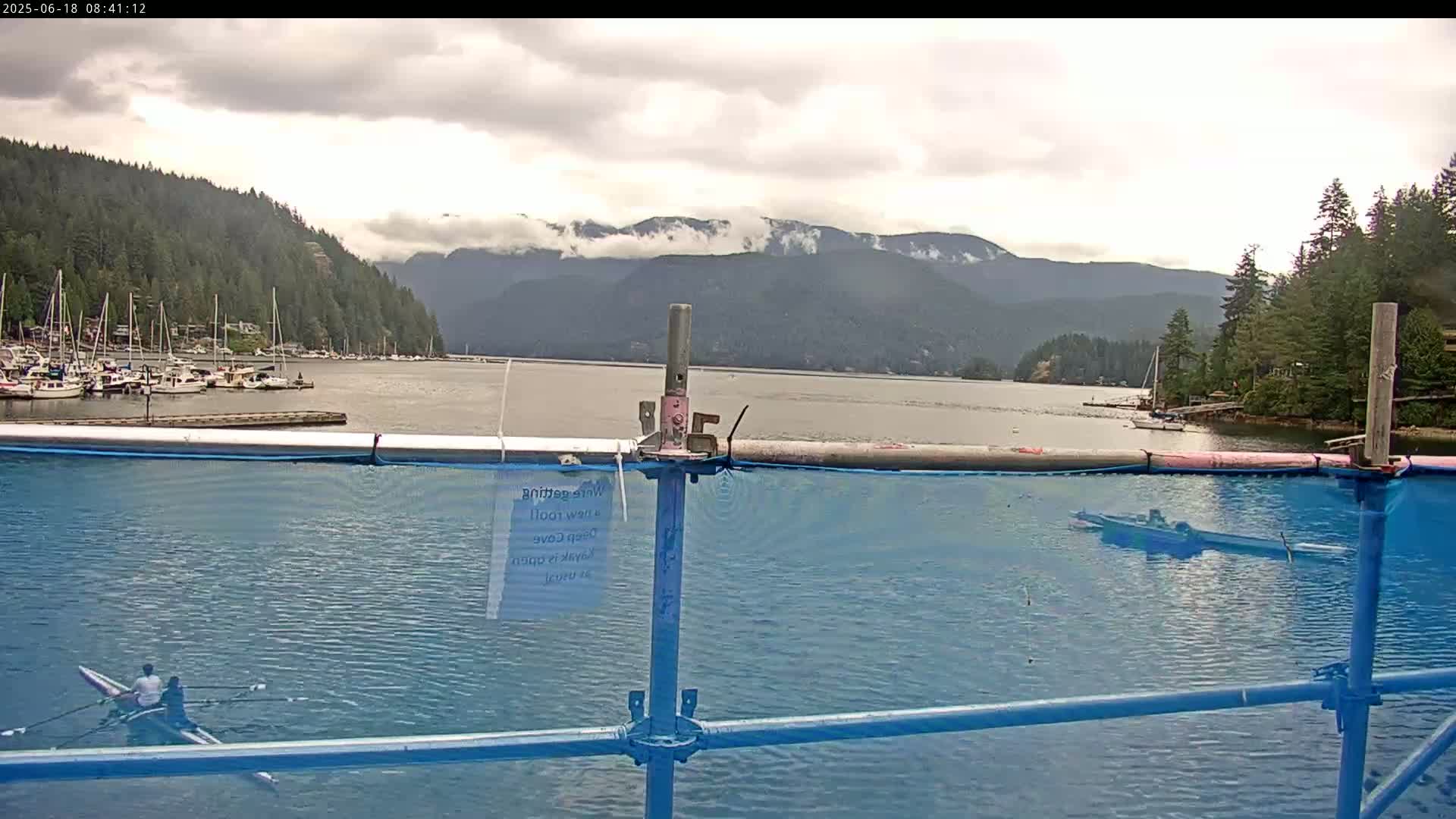 A calm bay with a marina, mountains in the background under a cloudy sky, is viewed from behind a blue mesh fence, with a rowing shell in the foreground.